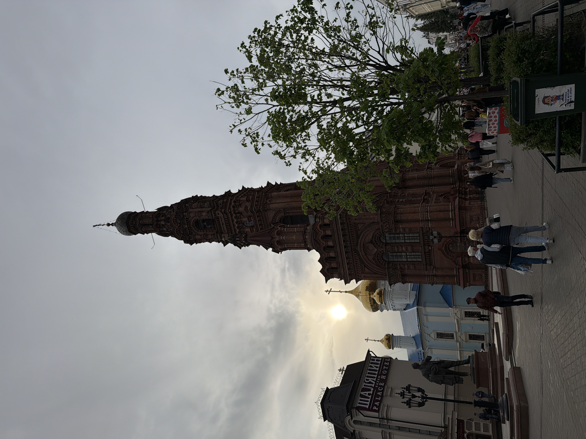 The image shows a street scene in Kazan, Russia. The scene features the leaning Soyembika Tower, and the golden domes of the Annunciation Cathedral, both part of the Kazan Kremlin complex. In the foreground is the Shalyapin Palace Hotel. People are walking along the pedestrian walkway. A statue and lampposts are also present. The sky is overcast.
