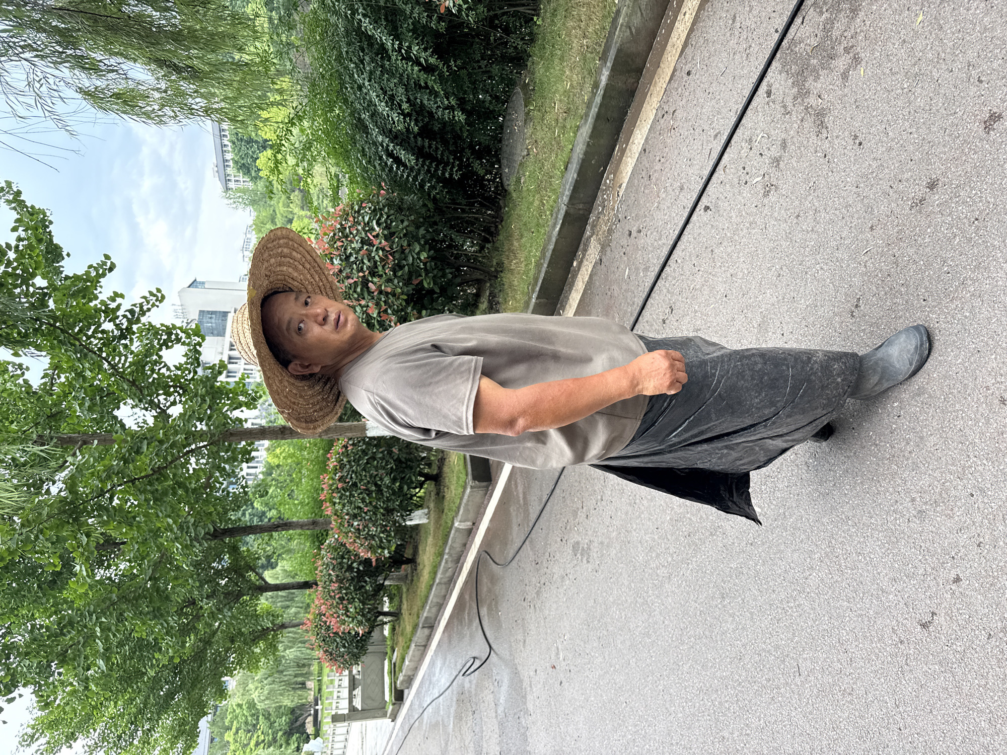 An Asian man wearing a straw hat, a short-sleeved t-shirt, dark trousers or a long skirt with rubber boots stands on a concrete paved area. The man is looking at the camera. Behind him, there is a landscape of trees and bushes along a white curb. A power cord lies on the pavement. The image was taken outdoors. The city is Jiangjin, China.