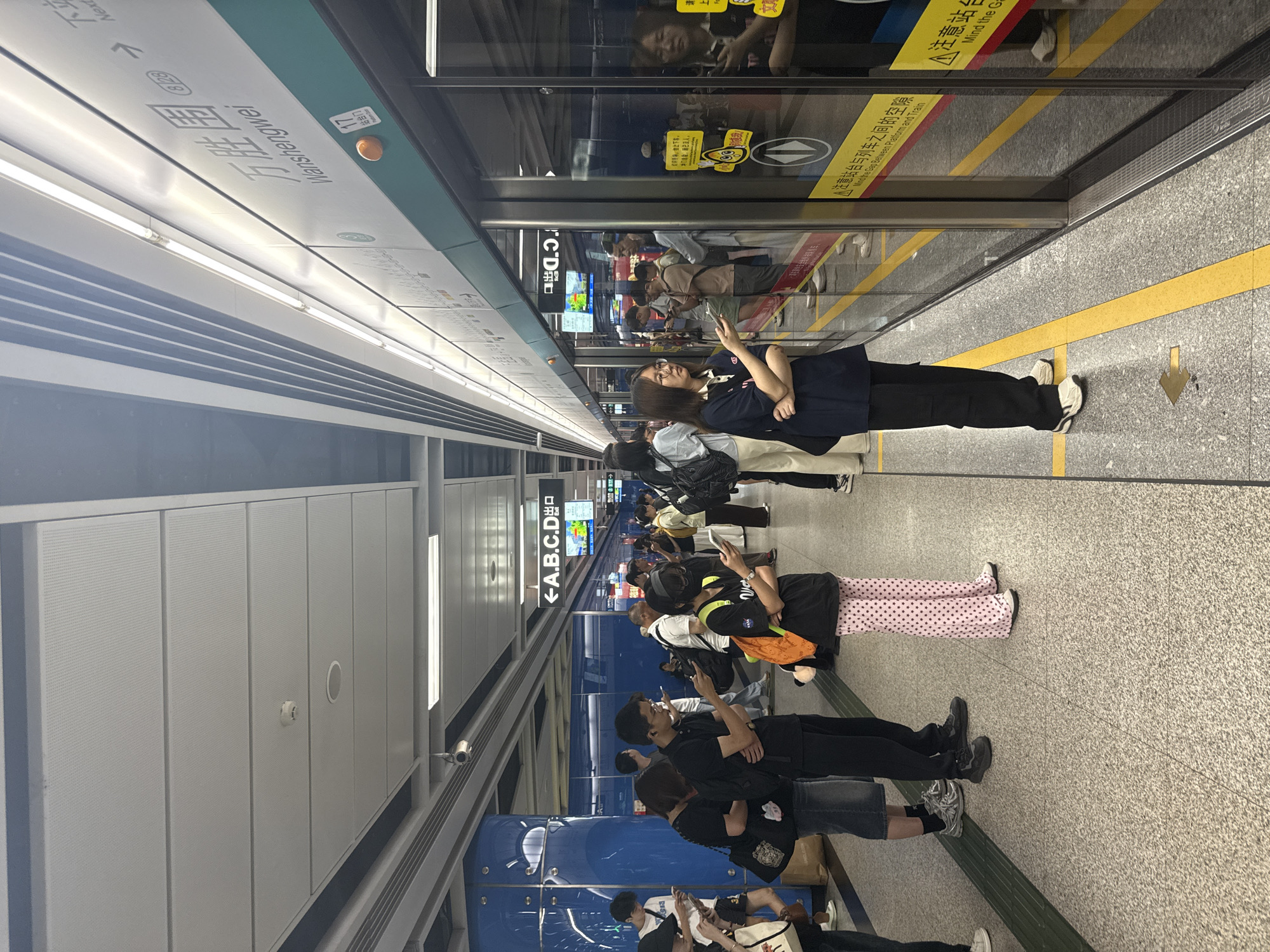This image captures a scene on a subway platform in Haizhu District, China. Several individuals are standing and waiting near the edge of the platform, facing a set of glass doors that likely lead to the train. The environment is a modern subway station with a grey, speckled floor marked with yellow lines. Overhead, a white ceiling with recessed lighting and a suspended track system is visible. Signs with Chinese characters and English text are present, including one that reads "Wanshengwei" and indicates "Next". People are dressed in casual attire, and many are looking at their phones. Some are holding bags or backpacks. The overall atmosphere suggests a typical urban commute.