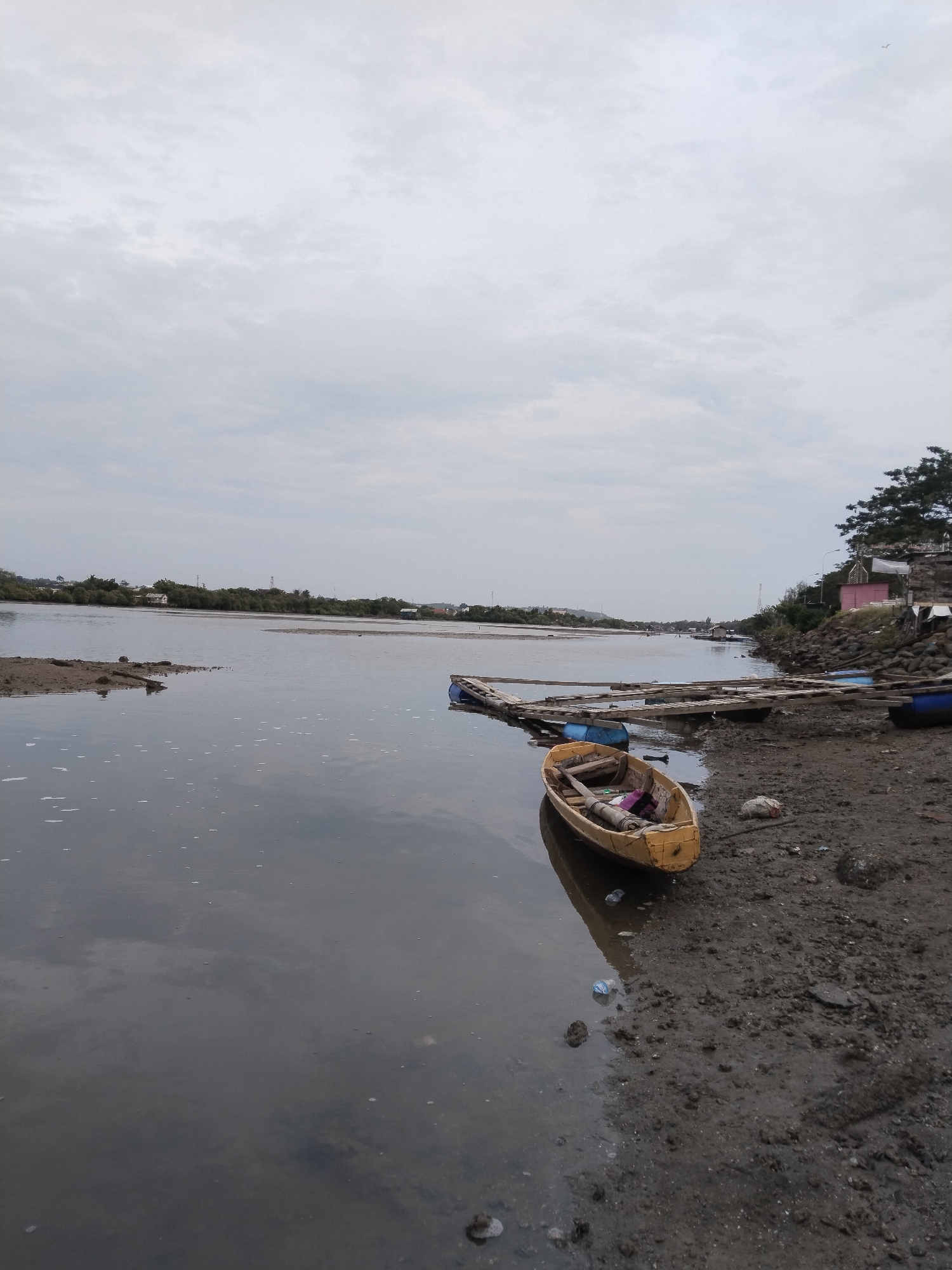 The image captures a waterfront scene in Lhokseumawe, Indonesia, under an overcast sky. In the foreground, a yellow wooden boat rests on the muddy shore, partially submerged in the calm water. The boat contains some items, including what appears to be fabric. Adjacent to the boat, a wooden walkway extends out onto the water, supported by blue barrels. Further back, the water stretches towards a distant tree-lined bank with several buildings visible. The shore itself is composed of dark mud and scattered debris, including plastic bottles. The water reflects the cloudy sky. To the right, the shoreline is rocky, with more structures, including a small pink building, nestled among trees. The overall impression is one of a quiet, somewhat utilitarian waterfront.