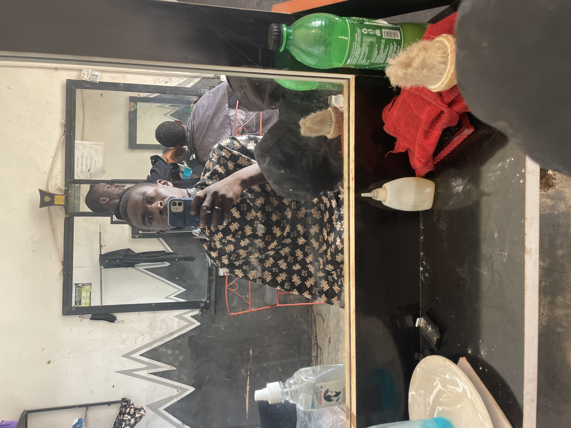 The image shows a man in a barbershop, reflected in a mirror. The man is holding a blue smartphone, presumably taking the photograph. He is wearing a patterned black and gold garment. The reflection shows the interior of the barbershop, with another person receiving a haircut in the background. The barbershop has a simple decor, with a wall painted with a black and white zigzag pattern. Various grooming items are visible on a shelf next to the mirror, including a green plastic bottle, a brush, a red cloth, and a white container. Based on the information, the location is identified as Durimi, Nigeria.