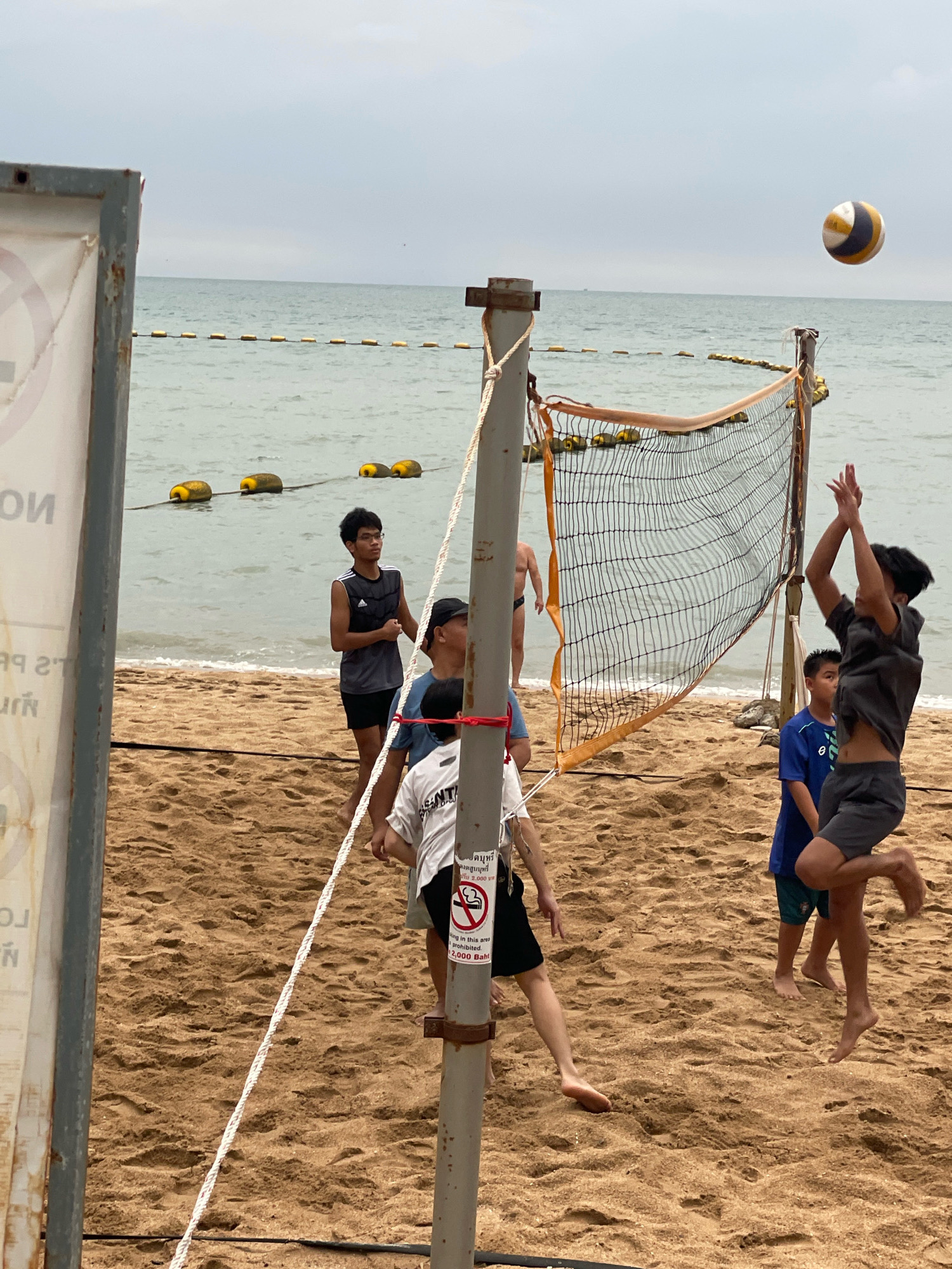 This image depicts a beach volleyball game in progress on a sandy beach in Pattaya City, Thailand. The scene is set against a backdrop of the sea and a cloudy sky. A volleyball net is strung across the sand, and several young men are actively participating in the game. One individual is mid-air, poised to hit the volleyball, which is visible in the upper right portion of the frame. Other players are positioned on the sand, observing the play or preparing for their next move. Yellow buoys are spaced out in the shallow water behind the players, marking a boundary in the sea. A signpost with text is visible on the left side of the frame, and another sign with a "no smoking" symbol and text in Thai and English is attached to the volleyball net pole. The sand is textured with footprints and indentations from the activity.