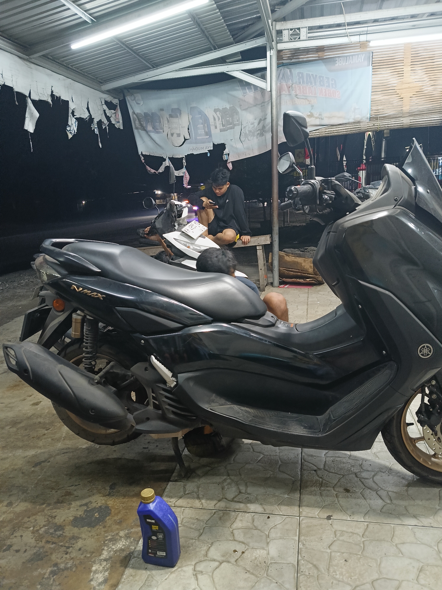 This image depicts a roadside motorcycle repair area or a makeshift workshop in Palangka Raya, Indonesia, likely at night due to the artificial lighting and dark background. A black Yamaha NMAX scooter is prominently featured in the foreground, parked on a tiled surface. To the left of the scooter, a blue bottle of motor oil with a yellow cap rests on the ground. In the background, a person is seated on a wooden bench, looking at a smartphone. Another person, possibly a child, is leaning against them. Behind the seated individuals, a white motorcycle is visible. The area is covered by a corrugated metal roof supported by metal poles, with torn fabric banners hanging from the roof's edge. The overall impression is of a functional, albeit somewhat unkempt, service area.