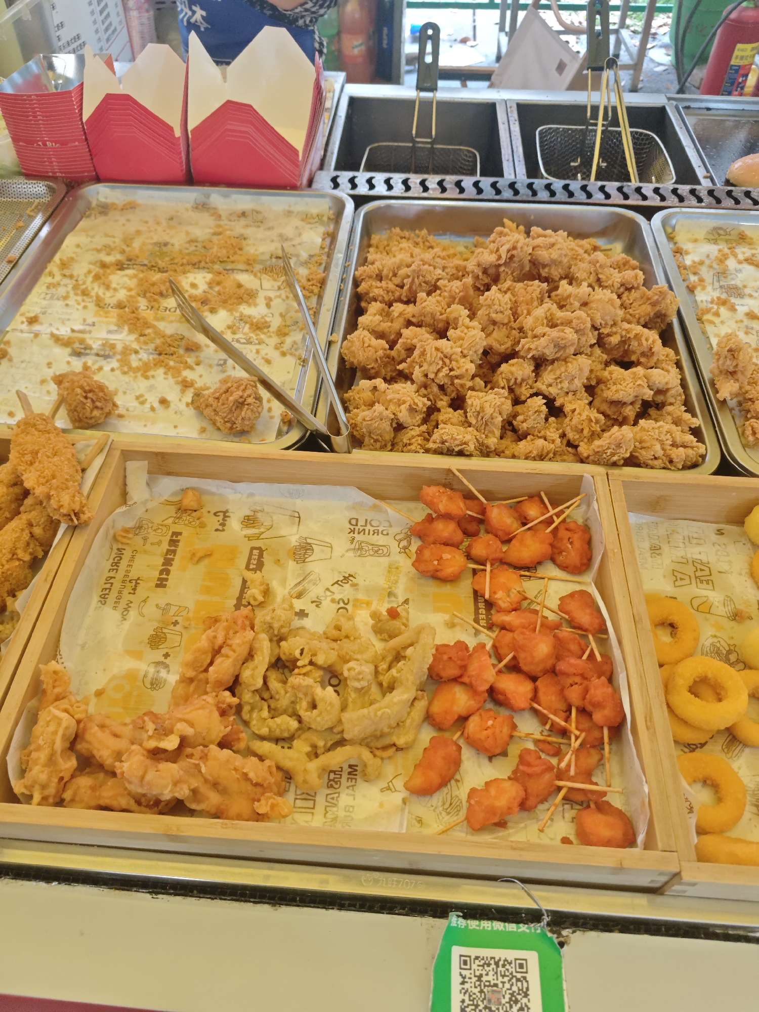 The image shows a street food vendor's display in an unidentified city. There are several trays and wooden containers filled with various types of fried food, including fried chicken pieces, fried chicken on skewers, fried bananas or similar, and fried onion rings. Utensils for serving are visible. The background shows frying equipment and possibly a customer. The items are being sold at a food stall in an outdoor environment. A QR code for payment is visible.