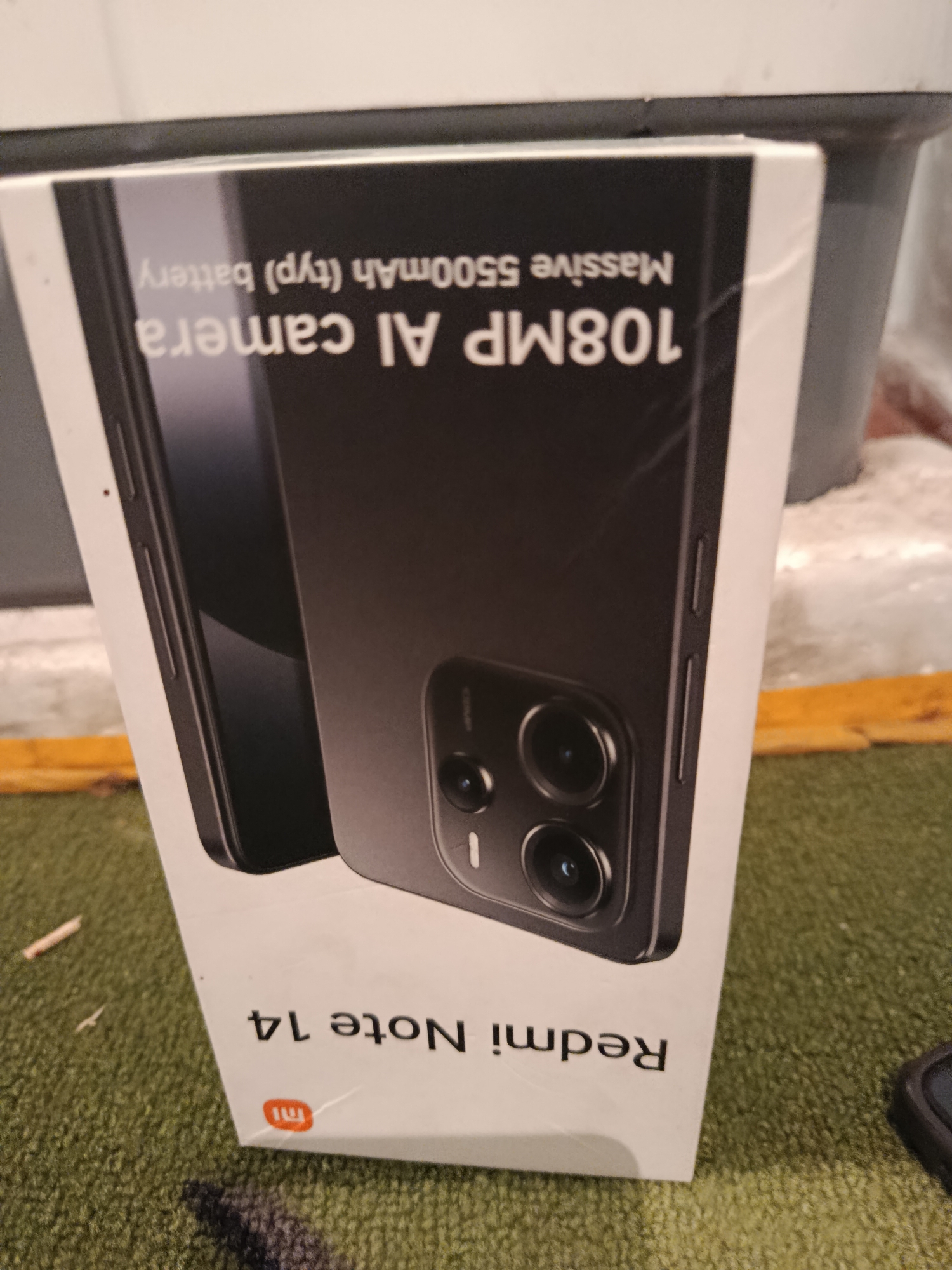 The image shows a close-up, angled view of a Redmi Note 14 smartphone box resting on a textured green surface. The box is white with black text and features an image of the phone itself, highlighting its "108MP AI camera" and "Massive 5500mAh (typ) battery." The brand logo, "mi," is visible in orange on the lower portion of the box. The lighting suggests an indoor environment, and the green surface appears to be a carpet or fabric. There are no people or discernable city landmarks present in the frame, making it impossible to determine its location within Jaŋı-Jol, Kyrgyzstan.