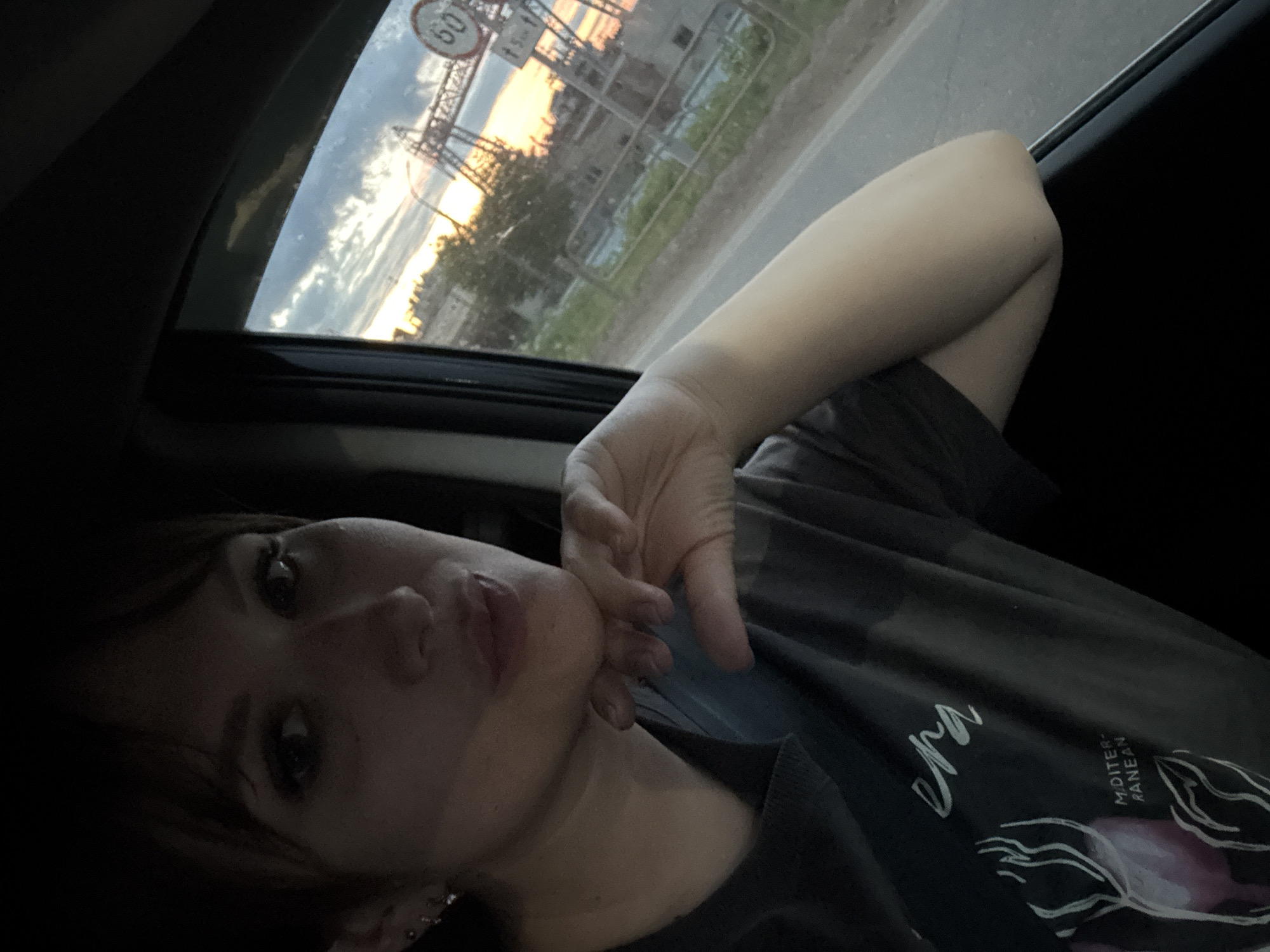 The image shows a person in a vehicle looking out the window. The location is likely Chaikovsky, Russia, as evidenced by the presence of a "60" speed limit sign and Cyrillic script in the window's reflection. The person has their face tilted towards the camera with their hand near their chin. They are wearing a dark t-shirt with a design. The background visible through the car window suggests a city scene with buildings and a colorful sunset.