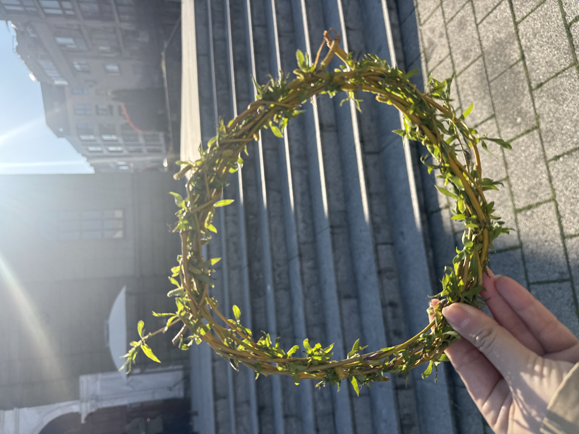 A close-up shot shows a hand holding a wreath made of twigs and small green leaves, likely willow branches, against a backdrop of an outdoor staircase. The staircase has multiple steps and is made of grey concrete. In the background, out of focus, there are buildings suggesting an urban environment. Based on the architecture, foliage, and specific architectural details, the location is likely Burgas, Bulgaria. The light suggests it is daytime. The hand is holding the wreath slightly to the right of the frame.