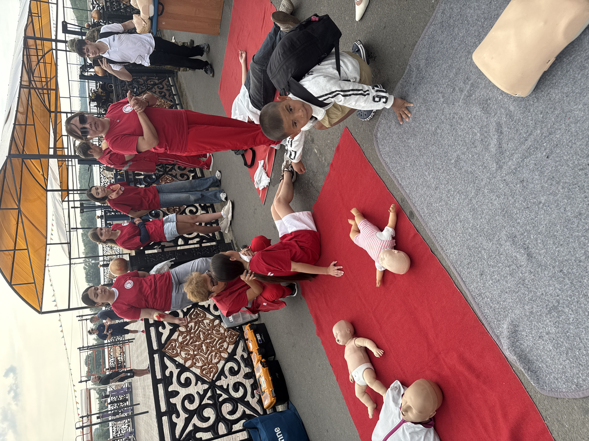 The image depicts an outdoor CPR training session. In the foreground, there are several CPR training mannequins, including infant and adult sizes, laid out on red and gray mats. A young boy wearing a white sweatshirt with the number "91" and a black backpack is kneeling on the gray mat, looking towards the camera. Behind him, an adult man is lying down on a red mat.
In the middle ground, a group of people, mostly women in red shirts, are participating in the training. One woman in red is kneeling and appears to be demonstrating a CPR technique on a young girl. Another woman in red, wearing sunglasses and a red headband, is standing and gesturing, possibly instructing the group. Other individuals, including a teenage girl and a boy in a white polo shirt, are also present, observing the training.
The background shows a paved area with decorative metal railings. Beyond that, there are some structures with yellow awnings and what appear to be exercise equipment. The sky is overcast.
The scene is likely taking place in a public outdoor space in the city of Pavlodar, Kazakhstan, given the context of a CPR training event. The presence of the Red Cross symbol on some of the red shirts suggests a connection to a humanitarian organization. The overall atmosphere appears to be educational and practical, focused on teaching life-saving skills.