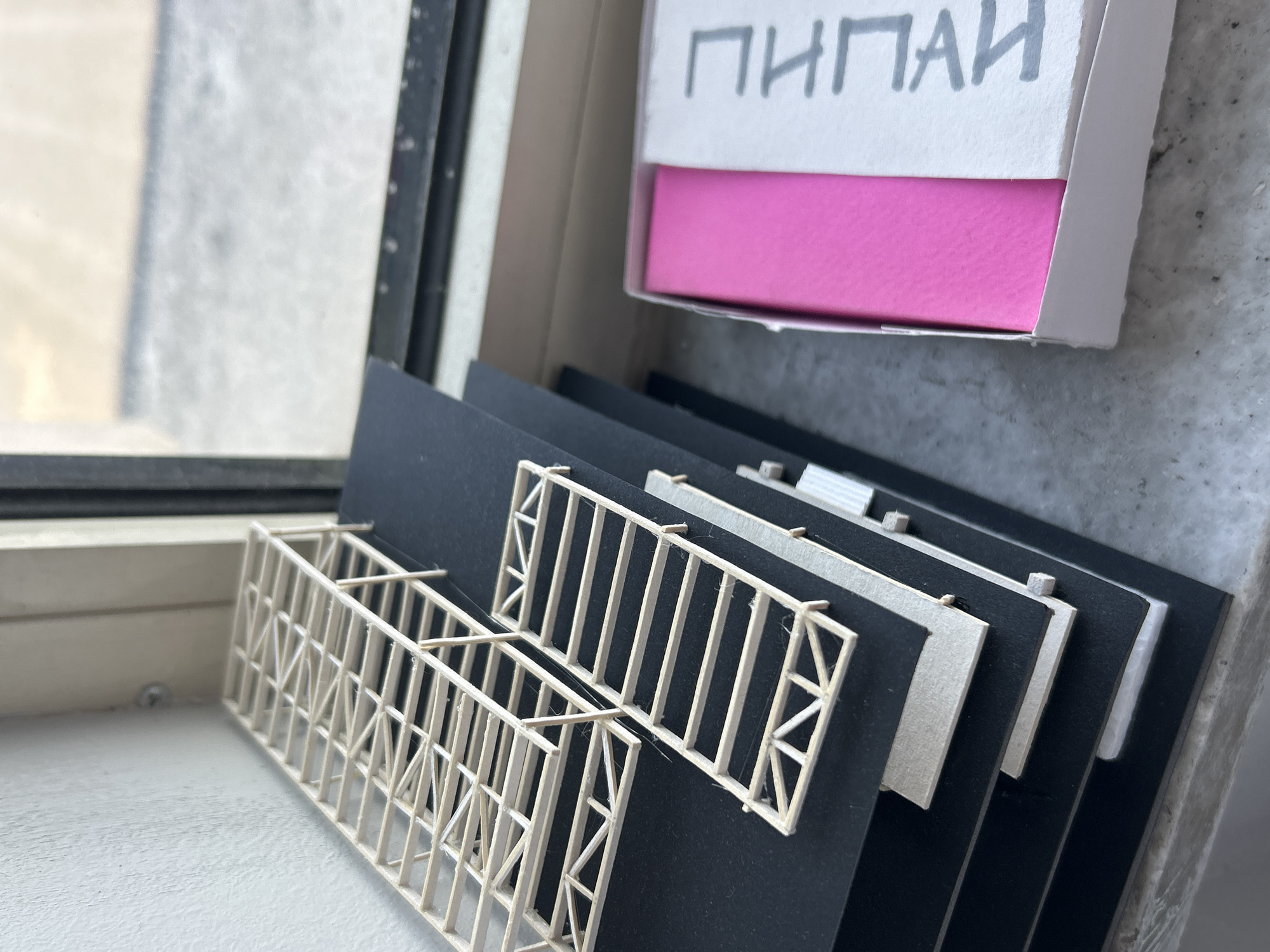 The image shows a close-up of several items on a windowsill. A white box with pink interior and Cyrillic script "ПИПAЙ" in dark grey letters sits to the right, partially obscuring a grey, textured wall. To the left of the box, a stack of black cards with attached white, lattice-like structures resembling architectural or industrial models rests on the windowsill. The windowsill is white and appears to be made of a synthetic material. The window itself is visible in the background, showing a blurry exterior. The environment is indoors, lit by natural light coming through the window. Sofia, Bulgaria is not discernible from the image.