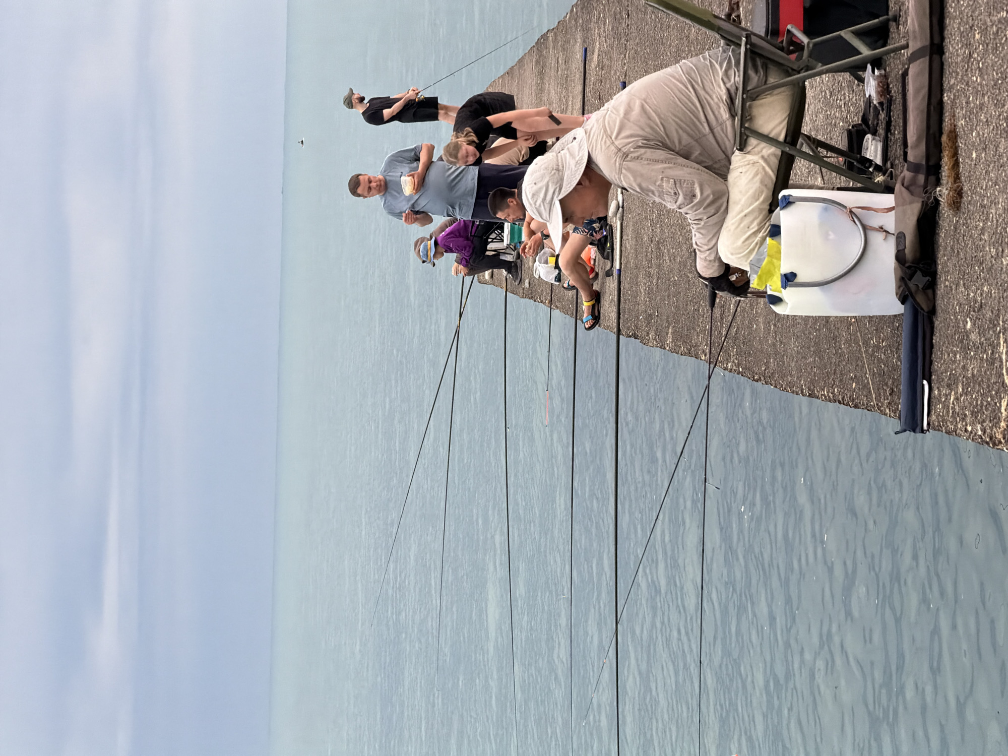 The image depicts a group of people fishing on a concrete pier extending into a calm, turquoise body of water, likely the Black Sea. The sky is overcast with a muted grey hue. The pier is made of concrete and appears somewhat weathered.
Several individuals are visible, engaged in fishing:
* In the foreground, a man wearing a light-colored long-sleeved shirt and a wide-brimmed hat is seated in a green folding chair. He is looking towards his fishing rod, which is positioned in front of him. A large white plastic container with blue straps sits next to his chair.
* Behind him and to his right, another person, a woman in a black dress, is leaning over, seemingly interacting with the fishing rods or the water.
* Further down the pier, a man in a light blue t-shirt and dark pants is standing and holding a small plastic container with food.
* To his left, a young person in a purple jacket and a blue hat is seated on a chair, also holding a fishing rod.
* Further back, a man in a black t-shirt and shorts stands with a fishing rod in his hand.
* Another person, a woman with dark hair, is visible in the middle ground, also appearing to be fishing.
Numerous fishing rods extend from the pier into the water, indicating active fishing. Fishing equipment such as tackle boxes and rod bags are present on the pier. The overall atmosphere suggests a casual recreational activity. Given the context of a pier on the Black Sea with numerous people fishing, this scene is consistent with a location in Sochi, Russia, known for its coastal attractions and recreational opportunities.