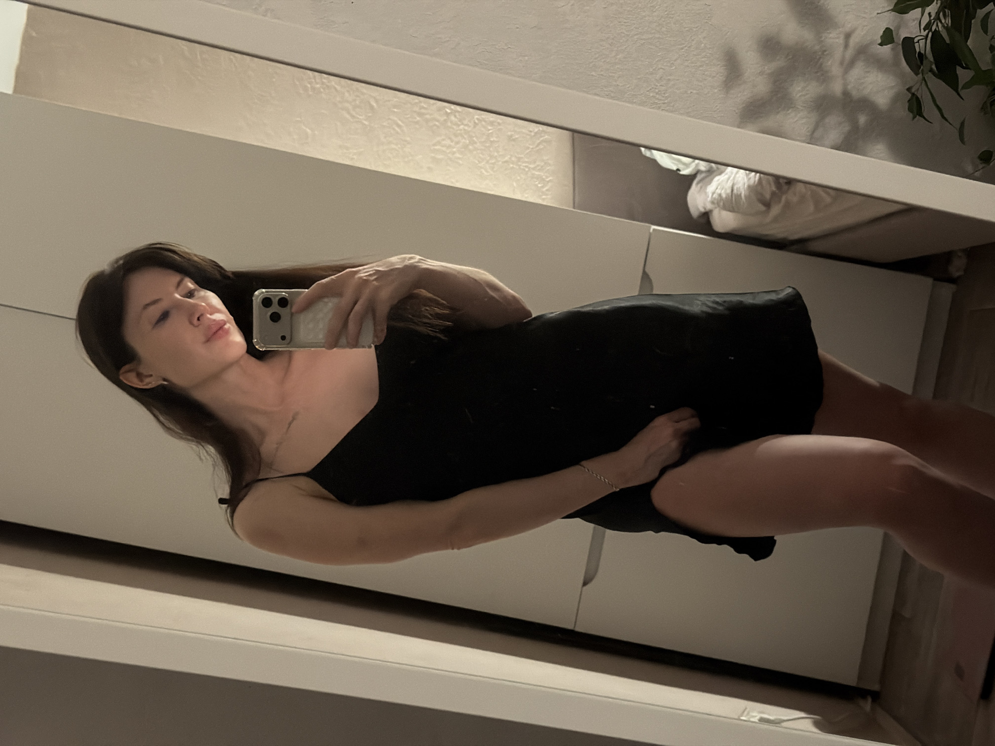 A woman with long brown hair, wearing a black slip dress, is taking a selfie in a mirror. The background includes a white wardrobe, a bed with white bedding, and a plant. The location is likely an interior setting such as an apartment. It is not possible to discern the city of Perm, Russia from this image.