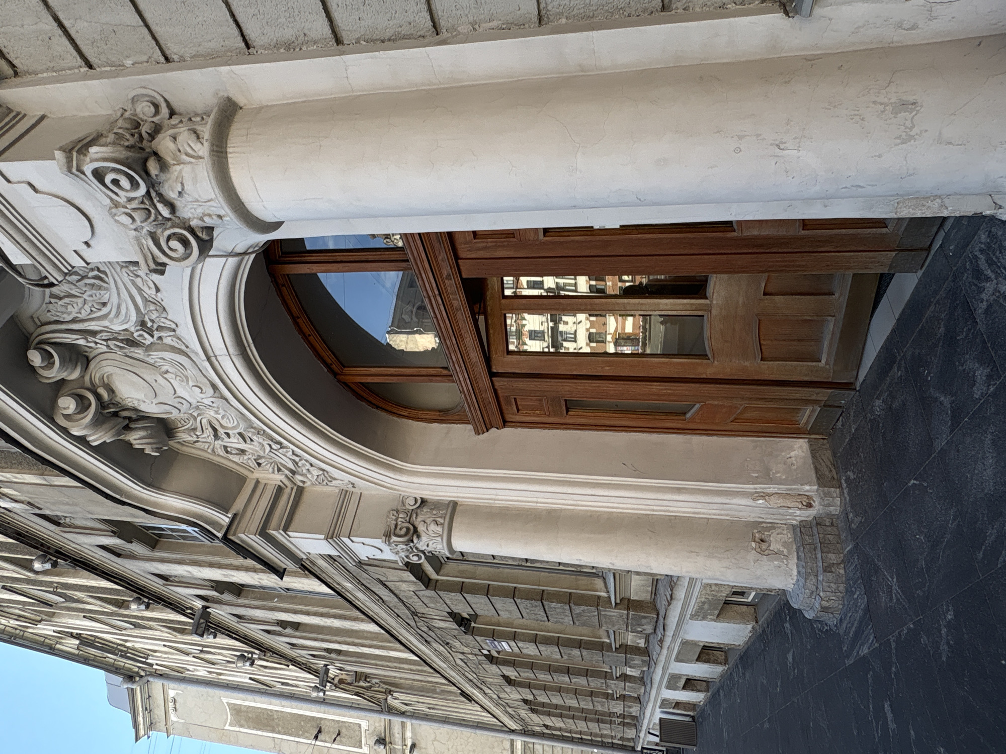 The image shows a close-up view of the entrance to a building, likely in Saint Petersburg, Russia. The architectural style is ornate, featuring a large wooden double door with glass panes, set within a stone archway adorned with decorative carvings and classical columns. The building facade is made of a textured stone or stucco material, with multiple windows visible along its length. The ground in front of the entrance is paved with dark, large-format tiles. The sky is clear and blue, indicating daytime. Reflections of buildings can be seen in the glass panes of the door. The overall impression is of an old, grand building in an urban setting.