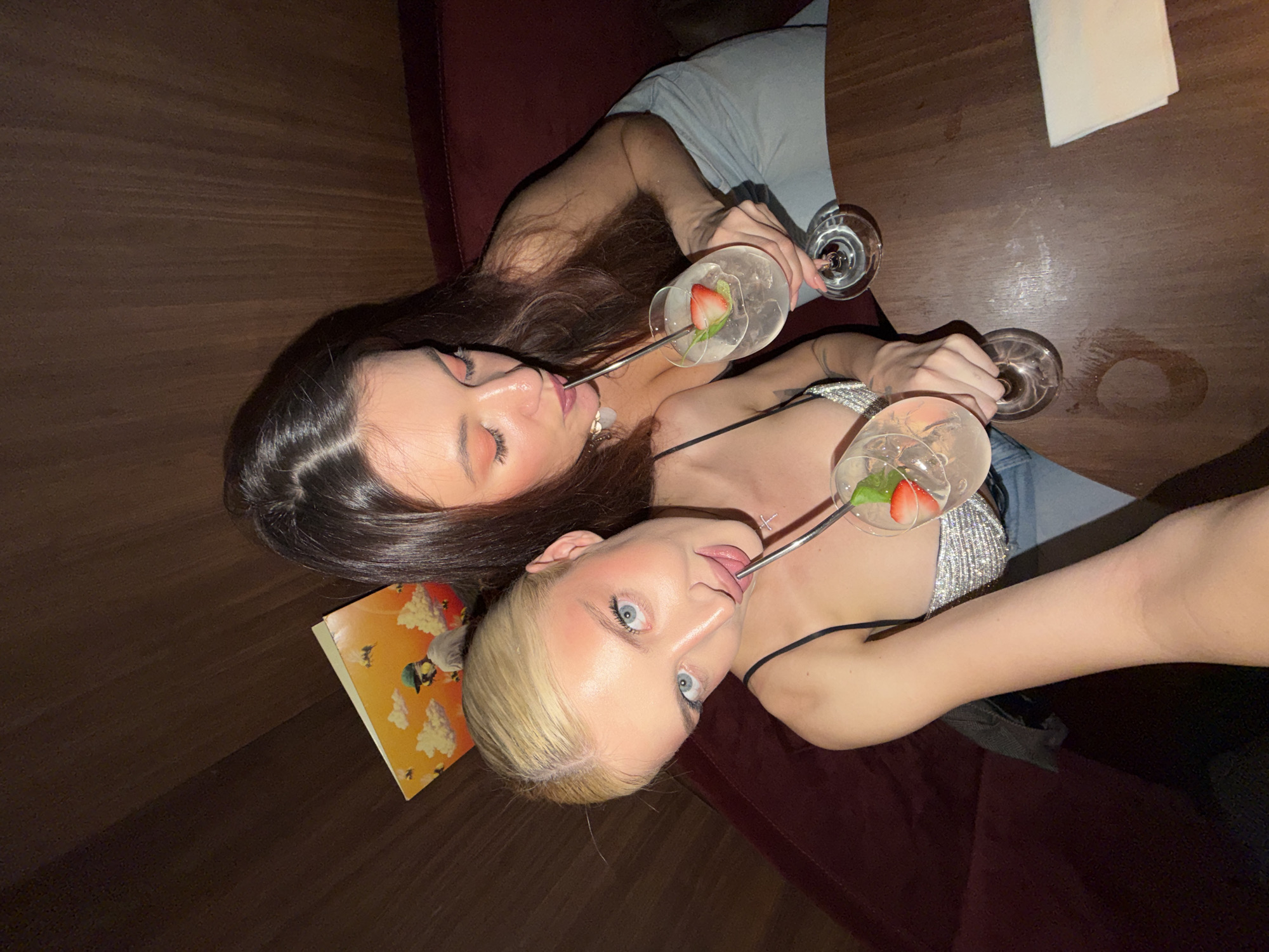 Two women are depicted from an elevated perspective, with one appearing to be capturing the image. The woman on the left has dark brown hair and is wearing a light-colored dress. The woman on the right has blonde hair and is wearing a sparkling top with thin straps. Both are holding stemware containing beverages, garnished with what appears to be strawberries, and are using metal straws. A small book with a yellow and orange cover is visible near the blonde woman. The setting seems to be a lounge or bar with a dark wooden table. One woman is sitting on a red velvet cushioned seating, while the other is sitting next to her. The precise location in the city is not discernible from the provided context.