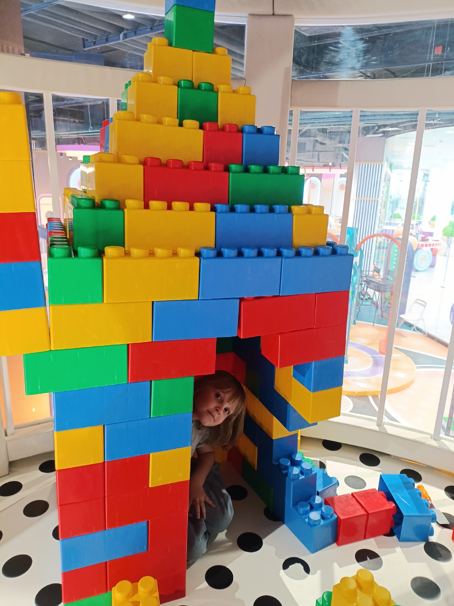 A close-up shot shows a young child playing inside a structure constructed from oversized Lego-like bricks. The structure, assembled with various colors (yellow, red, green, blue), forms a small enclosure. A child with light hair peeks out from inside the structure. The floor is white with black polka dots. Outside the structure, more building bricks are scattered around, and a window provides a view of an indoor play area with colorful play structures and equipment. The location is Unknown, Unknown.
