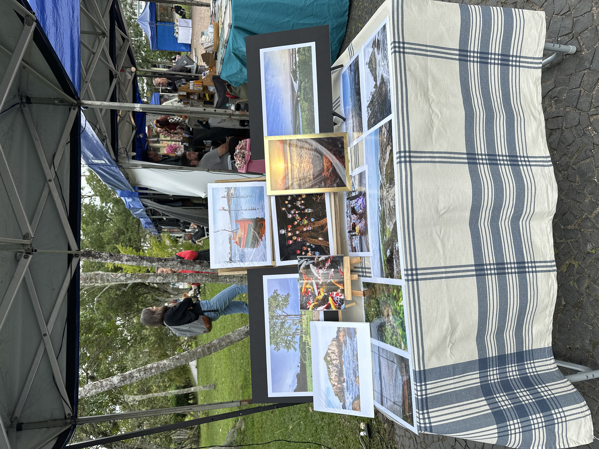The image shows an outdoor market stall, likely in Florianópolis, Brazil, given the context of a market and the style of goods displayed. A table covered with a blue and white plaid tablecloth is laden with framed photographs, predominantly featuring seascapes, landscapes, and a few close-ups of colorful fish (koi). Behind the table, a tent structure provides shade. In the background, several people are visible, some browsing or interacting at other stalls, and one person is walking away from the camera wearing a backpack and jeans. The environment appears to be a park or a public space with trees and paved ground. The photographs on display are the main subjects, showcasing various scenic views and artistic compositions.