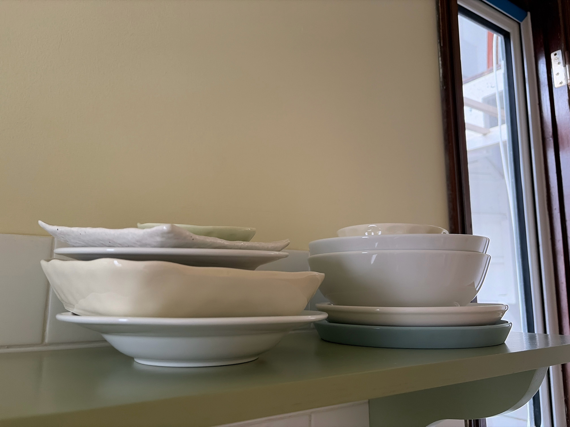 A stack of ceramic dishes is displayed on a shelf. The dishes are white and light yellow in color. The shelf is painted a light green color and is set against a beige wall. To the right of the shelf, a window with dark wooden trim is visible. The scene is indoors. There are no people or animals present.