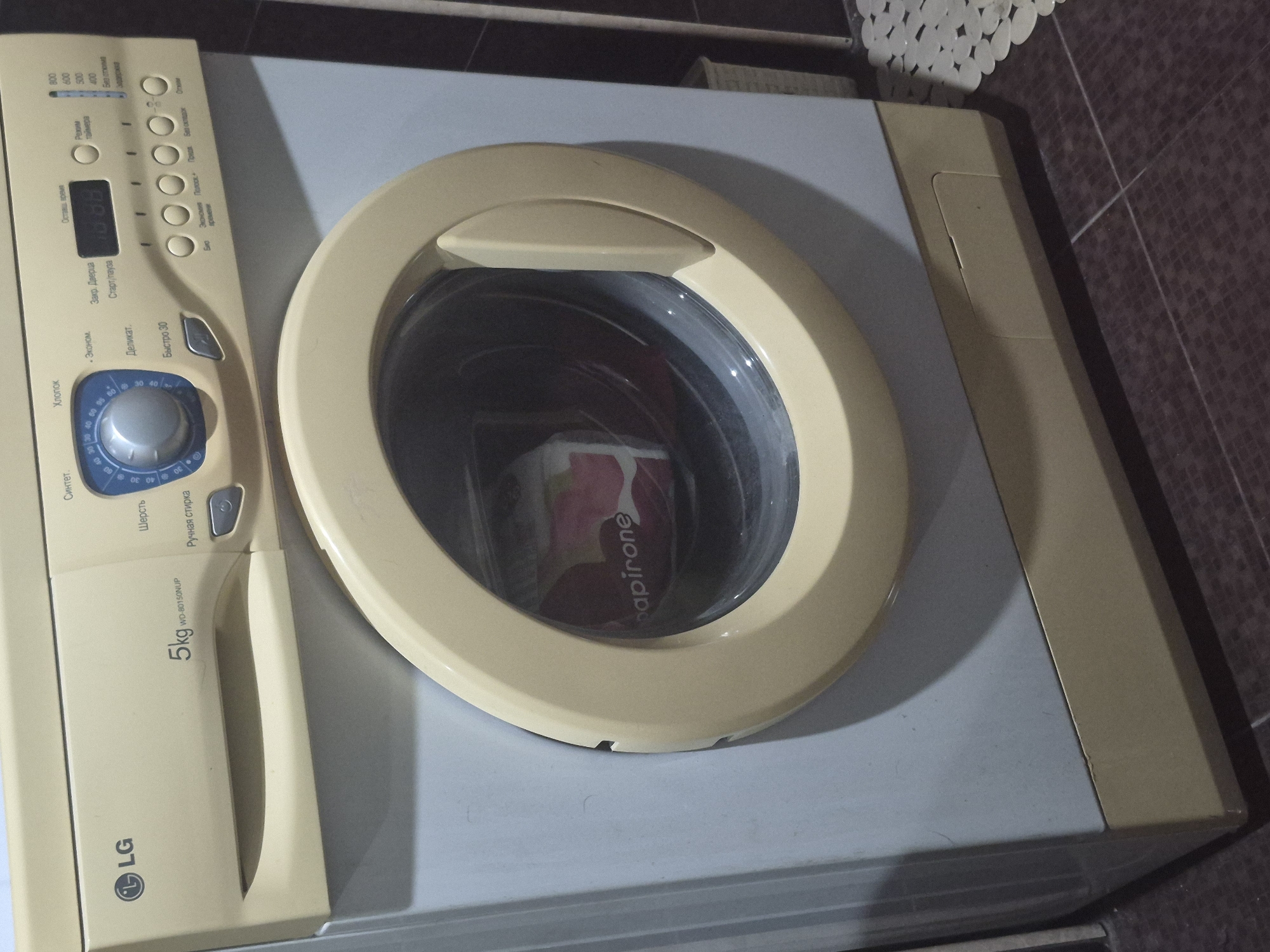 The image displays a cream-colored LG front-loading washing machine, model WD-80150NUP, with a 5kg capacity, in an indoor setting. The machine's control panel features a large program selection dial and several buttons, along with a digital display showing "1:38", likely indicating remaining cycle time.
Visible text on the control panel, primarily in Russian, includes wash programs such as "Хлопок" (Cotton), "Синтет." (Synthetics), "Шерсть" (Wool), "Ручная стирка" (Hand wash), "Эконом" (Economy), "Деликат." (Delicate), and "Быстро 30" (Quick 30). Spin speed options like "800", "600", "500", "400", and "Без отжима" (No spin) are also present. Other buttons or indicators include "Закр. Дверца" (Door locked), "Старт/пауза" (Start/Pause), "Режим таймера" (Timer mode), "Экономия времени" (Time saving), "Полоск." (Rinse), "Предв." (Pre-wash), "Без складок" (Crease care), "Отжим" (Spin), and "Био" (Bio).
Inside the circular glass door of the washing machine, some laundry, possibly a red or pink piece of fabric with the word "papirone" printed on it, is visible. The machine has a light gray top surface. The washing machine is placed against a wall with dark brown tiles, and a white, textured mat or cover is partially visible in the upper right corner of the frame. The scene suggests a domestic laundry area. No people are visible. The time of day is not discernible, but the lighting appears to be artificial, consistent with an indoor setting. The location context is Yerevan, Armenia.