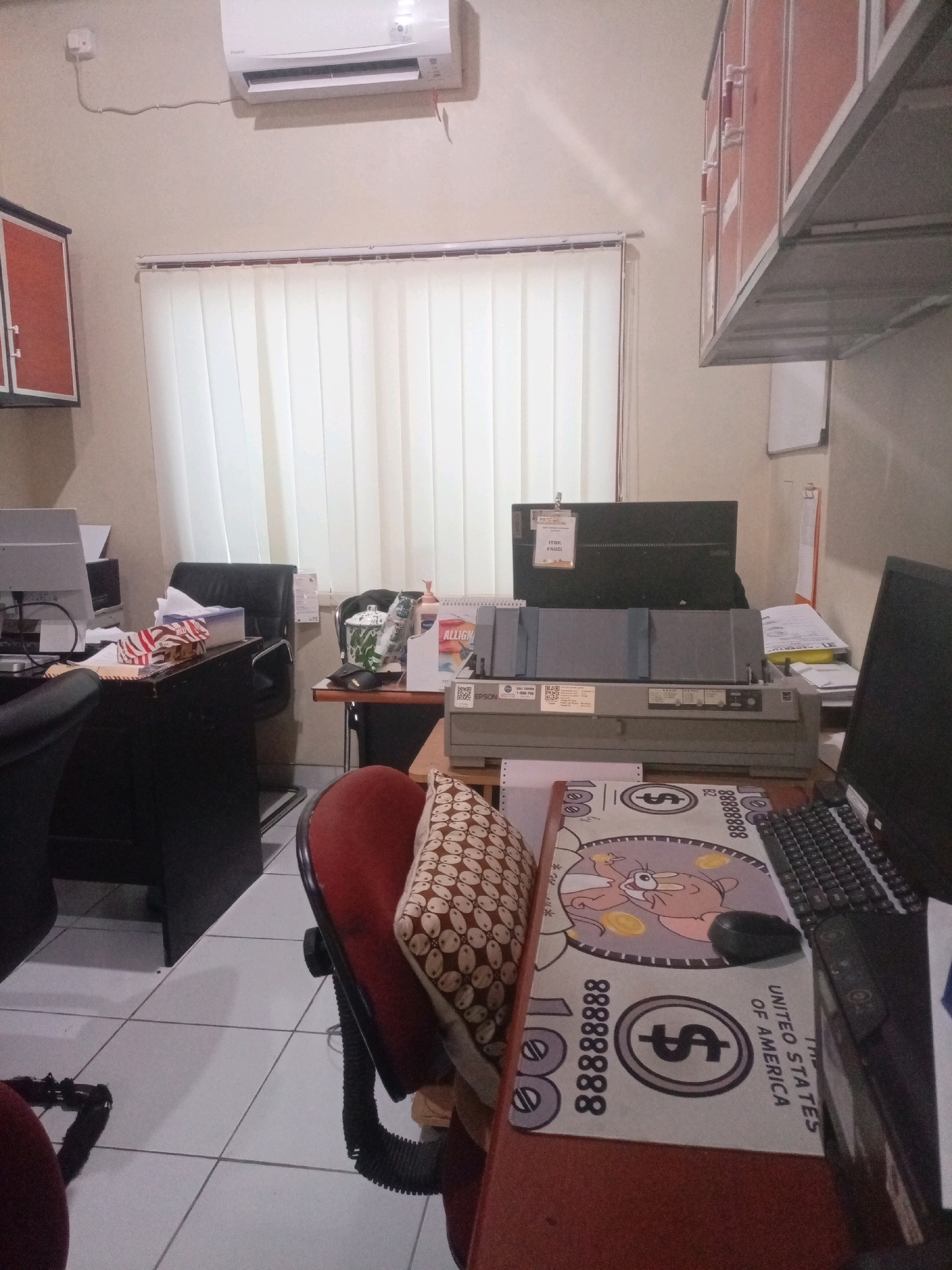 This image depicts an office interior located in Sumbersari, Indonesia. The room appears to be a workspace, possibly an administrative office or a cubicle. The walls are painted a neutral color, and vertical blinds cover a large window.
Dominating the scene is a large, grey dot matrix printer situated on a desk. A computer monitor, a keyboard, and a mouse are also visible on the desk, suggesting active computer use. To the left, a black office chair is positioned, indicating someone has been or will be working at the desk. A tissue box and some papers are also on the desk.
In the background, another desk is visible with a black office chair. Above this desk, a large air conditioning unit is mounted on the wall. There are also cabinets with orange and red fronts mounted on the wall.
On another desk in the foreground, a red office chair is present, and a pillow with a patterned design is placed on it. A mousepad with a cartoon character and dollar bill designs is also on this desk, along with a portion of a laptop.
The overall impression is that of a functional, albeit somewhat cluttered, office environment. The presence of office equipment and furniture suggests a place of work.