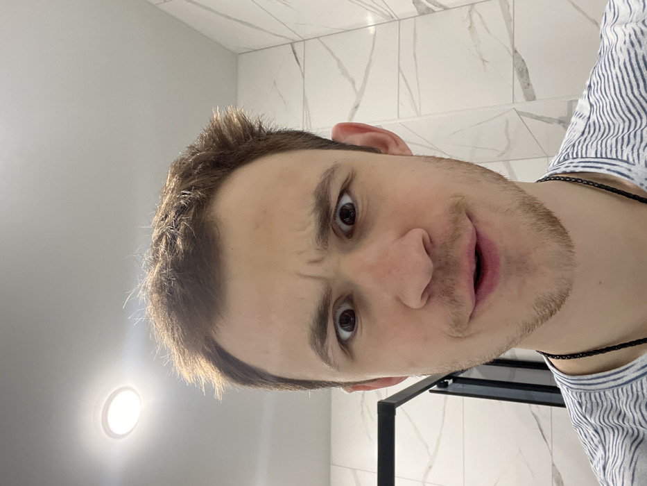 A close-up image captures a young adult male in an indoor setting, purportedly in the city of Tugurgoi, Russia. The subject has light brown, disheveled hair, fair skin, and light stubble on his chin and jawline. He is wearing a white and light blue horizontally striped t-shirt and a thin black cord necklace. His eyes are wide open and directed at the viewer, with his eyebrows furrowed, creating a puzzled or surprised expression. His mouth is slightly open. The background features light-colored, possibly white, rectangular wall tiles with subtle grey veining. A circular recessed ceiling light is visible in the upper left quadrant, and a dark, vertical structural element is partially discernible on the left side of the frame.