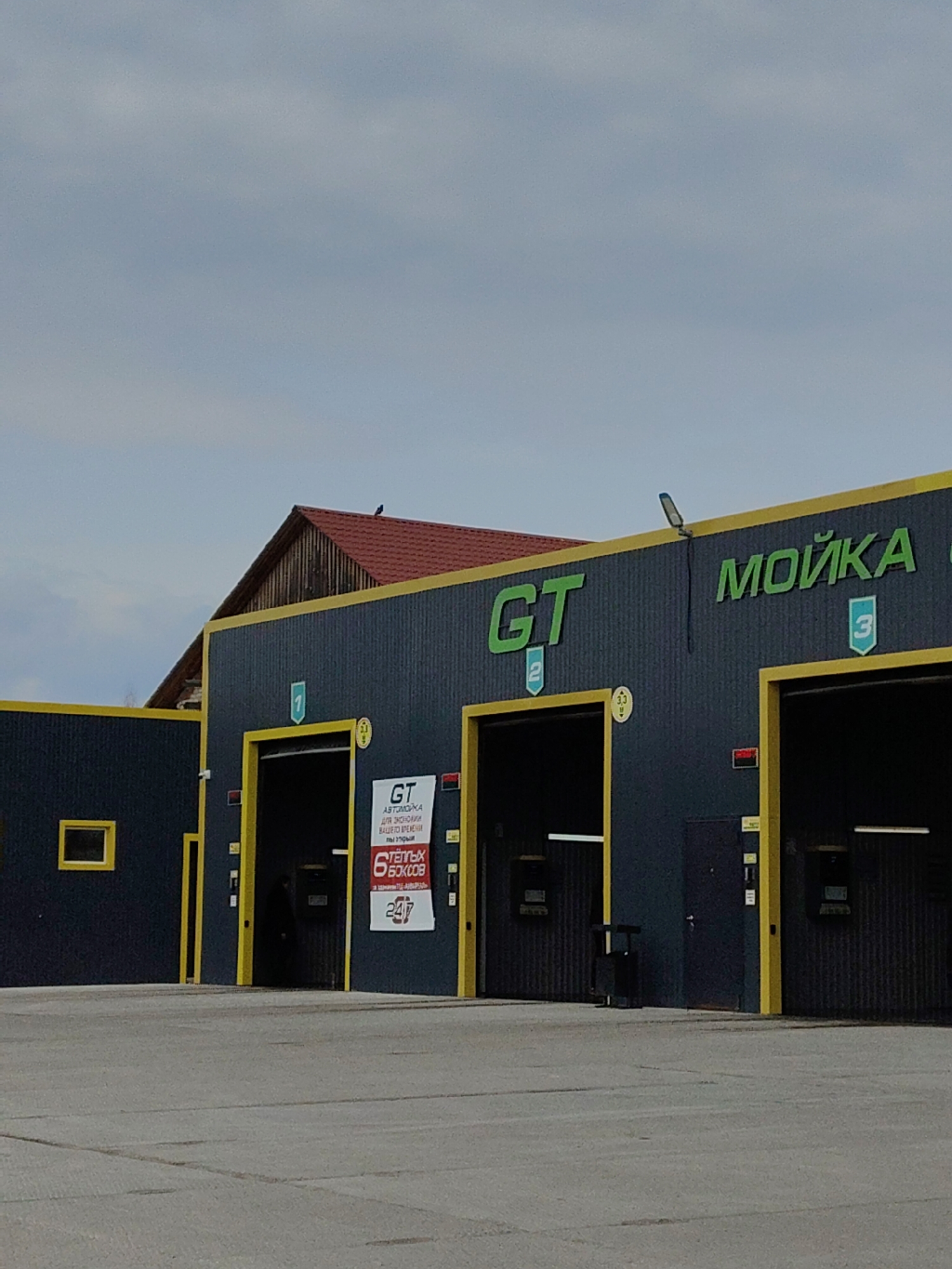 The image shows a car wash facility located in the city of Unknown, Unknown. The building is dark grey with yellow trim around the doorways and roofline. Each bay has a number above it, with the numbers 1, 2, and 3 visible. The words "GT" and "МОЙКА" are prominently displayed on the building in green letters. A sign near the first bay advertises "GT Автомойка" and "6 ТЁПЛЫХ БОКСОВ" (6 warm boxes) and "24/7". A small red roof is visible behind the car wash. A bird is sitting on the edge of the roof. The sky is overcast. The ground is paved with concrete.