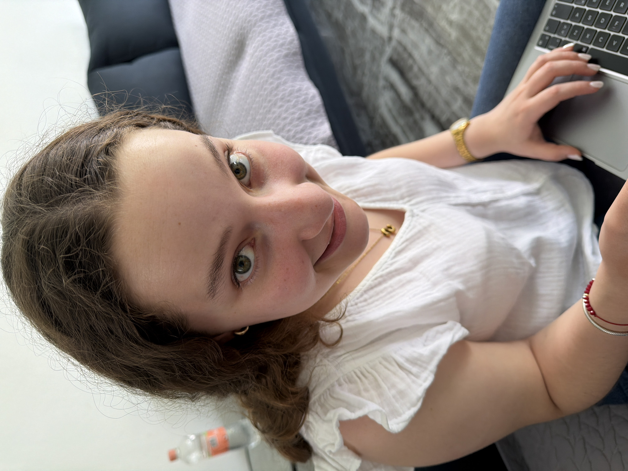 A woman is shown from a high-angle, close-up perspective. She is seated and using a laptop, her hand visible on the keyboard. She wears a white shirt and a gold watch and necklace. A red beaded bracelet adorns her wrist. Her light skin is visible. Part of a sofa, a pillow, a blanket, and a water bottle are visible in the background. The location of the scene is not discernible as being in the city of Puebla, Mexico.