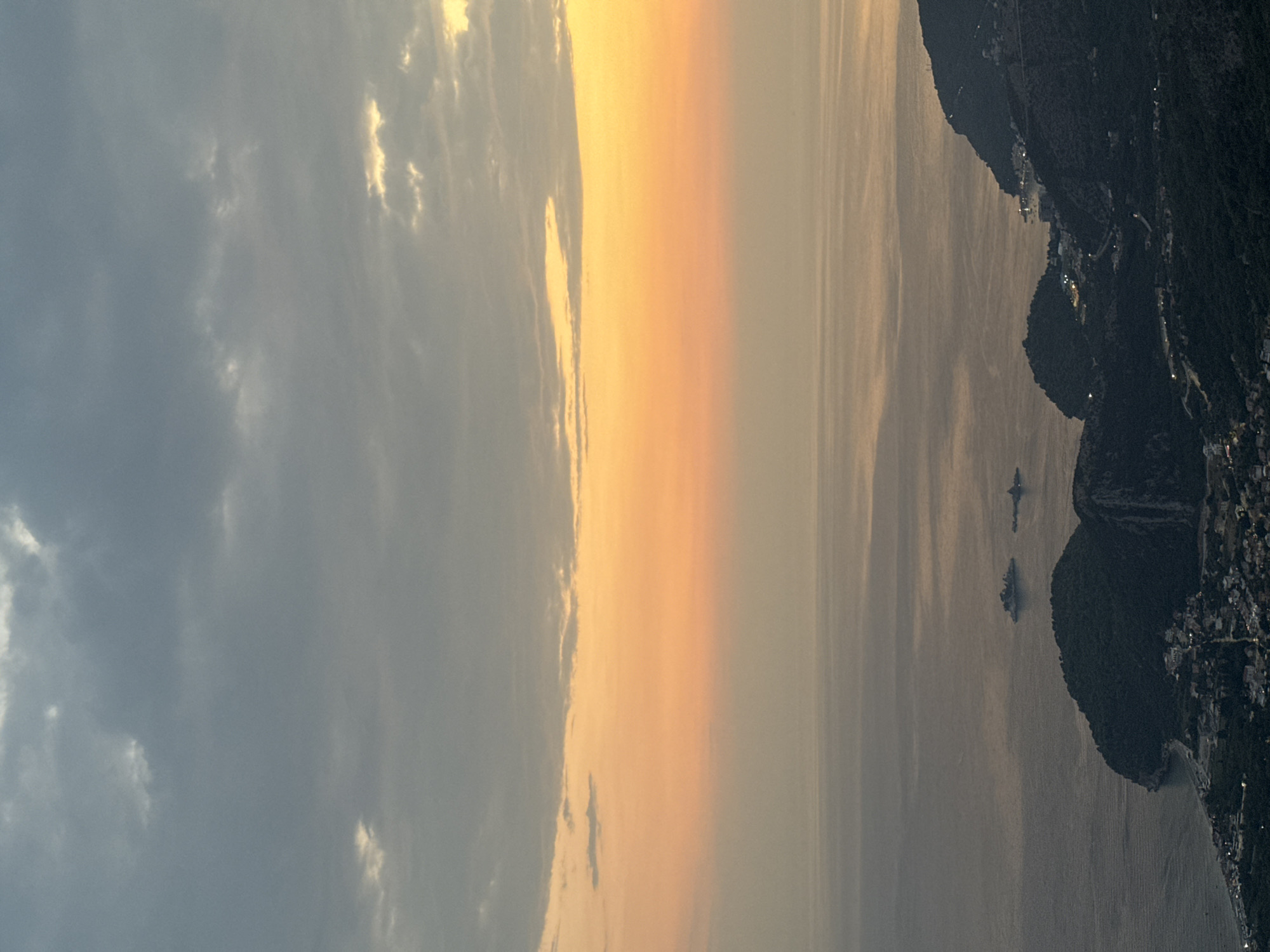 An aerial, eye-level, wide-angle, long shot captures a dusky, hazy seascape with a village nestled in the hills, a portion of the Adriatic Sea visible, and a gradient sky transitioning from orange to gray. The scene is devoid of people. Two small, dark, rocky islands, possibly Sveti Stefan and Sveti Nikola, are visible in the water, with a larger ship to their left. The coastline is rugged, with steep, tree-covered hills plunging into the sea, dotted with buildings that appear to be illuminated by lights. The sky displays a layer of dark, diffused clouds above a softer layer of pale orange and yellow, indicative of a sunset or sunrise. The water is calm and reflects the sky's colors. The location is identified as near Gluhi Do, Montenegro.