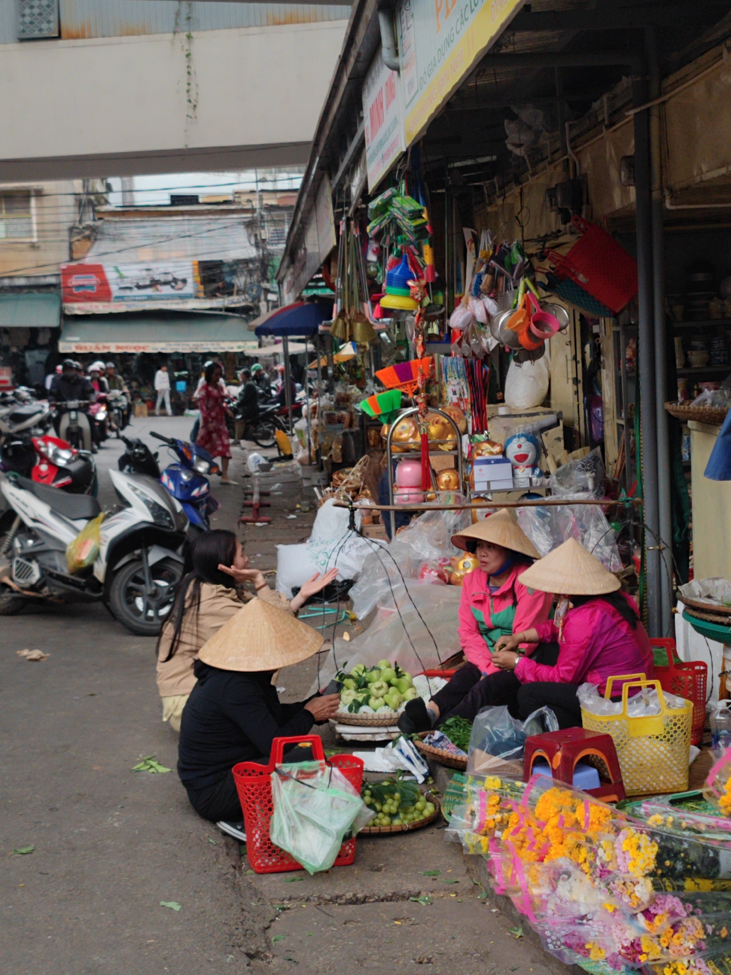 The image depicts a street scene in Da Lat, Vietnam. Several women wearing traditional conical hats are seated on the ground, engaged in selling fruits and flowers. The women are positioned in front of a shop selling various items. Motorcycles are parked along the roadside, and other pedestrians are visible in the background.