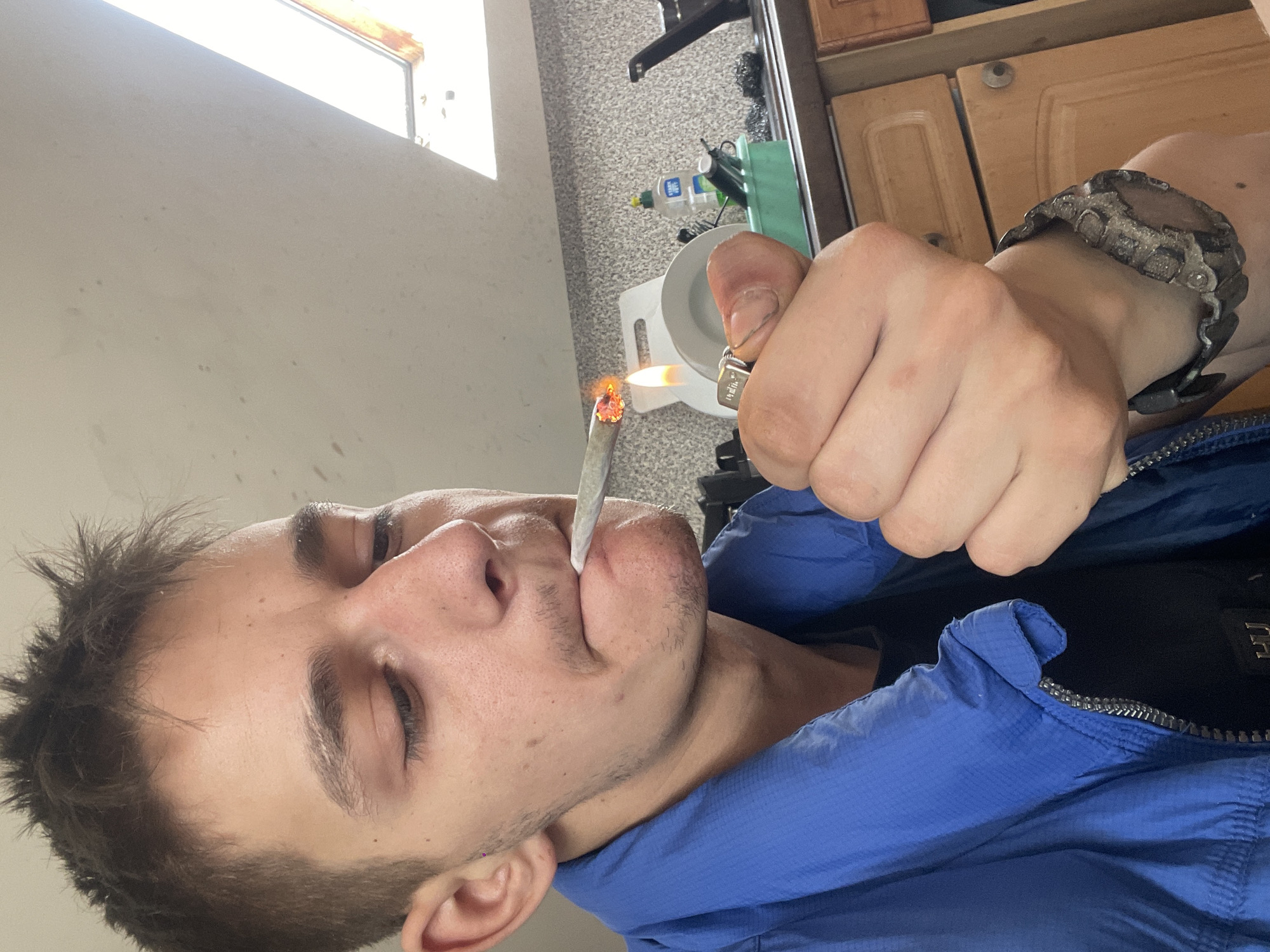 A young man with short, spiky brown hair and light skin is shown from the chest up. He is wearing a blue jacket over a black shirt. His eyes are closed, and he is holding a lit cigarette to his lips with his right hand. His left hand is holding a lighter to the tip of the cigarette, which is glowing orange. The background shows a kitchen counter with cabinets, a sink, and a bottle of dish soap. There is also a skylight in the ceiling. The image appears to have been taken indoors.