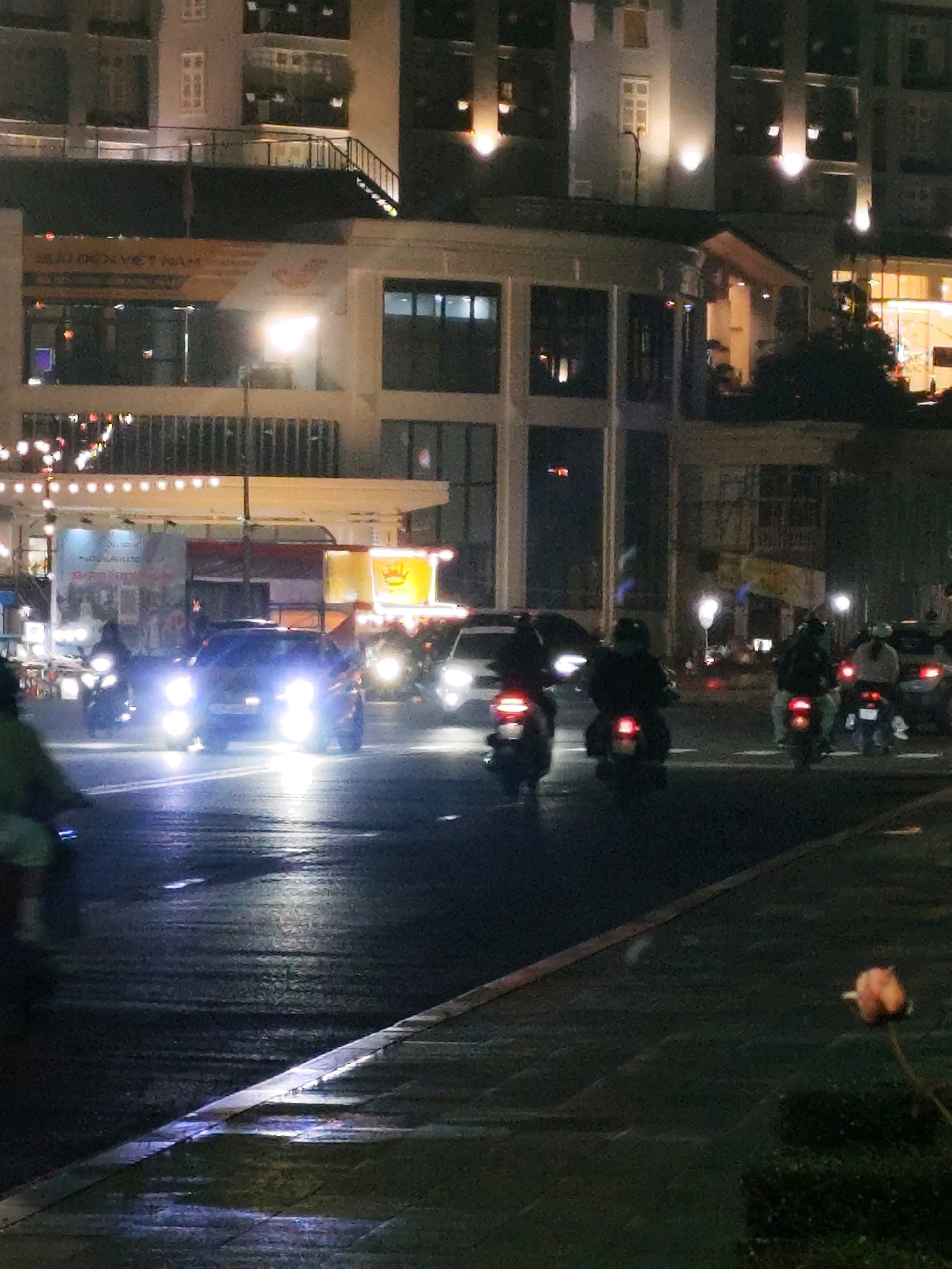 This is a nighttime photograph of a city street in Da Lat, Vietnam. The street is wet, reflecting the bright headlights of several vehicles and the neon lights of businesses. In the foreground, a car with its headlights on is driving down the road. Behind it, several motorcycles are also visible. The buildings lining the street are illuminated by indoor lights and exterior lighting. The overall impression is one of a bustling urban environment at night, with traffic and commercial activity.