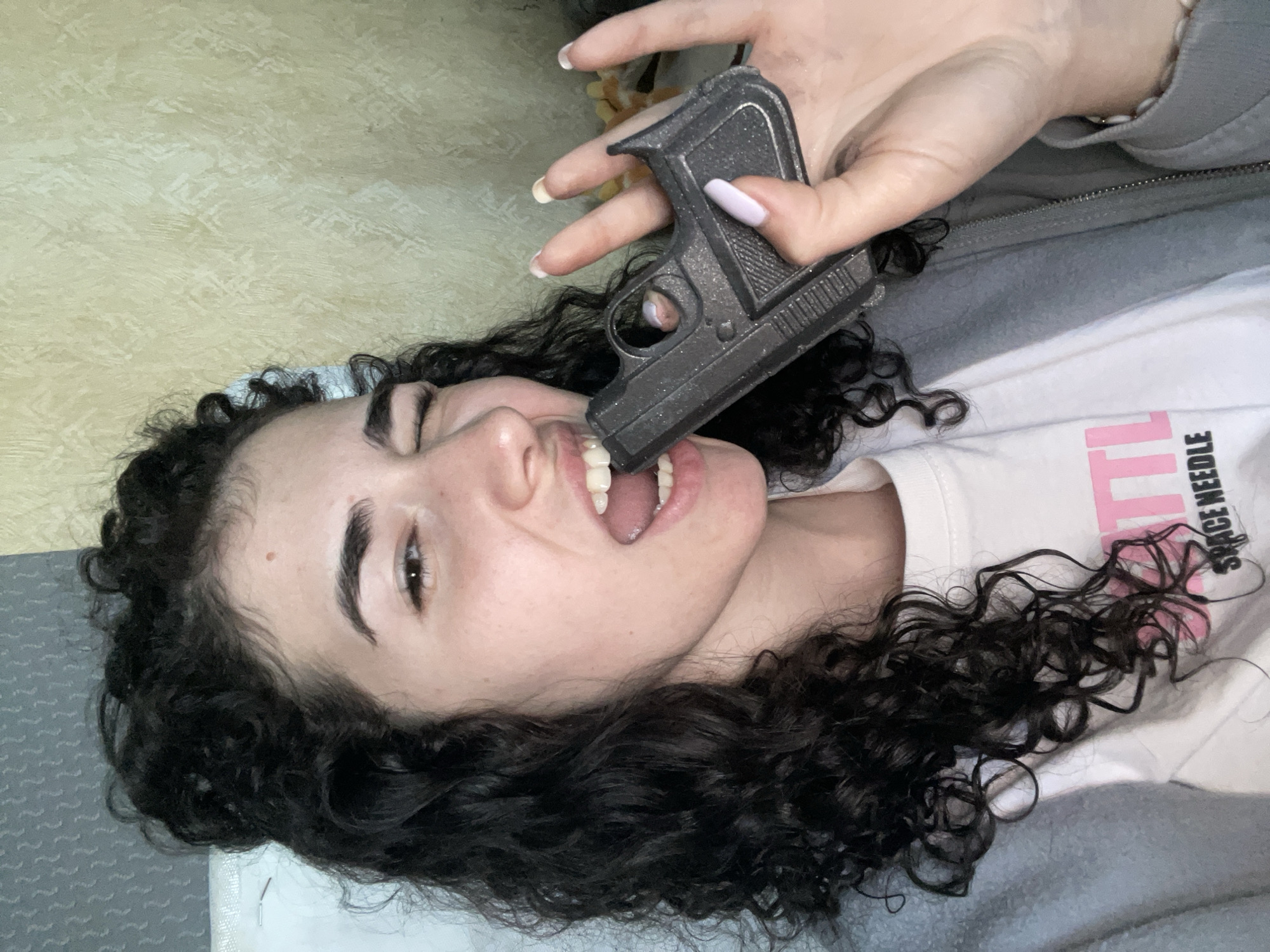 A woman with curly black hair is lying down. Her head is tilted back, and she is holding a toy gun in her mouth. The gun is gray and appears to be made of metal or plastic. She is wearing a white t-shirt and a gray jacket. Her fingernails are long and painted light pink. The background is a light yellow wall. The image does not contain information related to the location in the city of Sofia, Bulgaria.