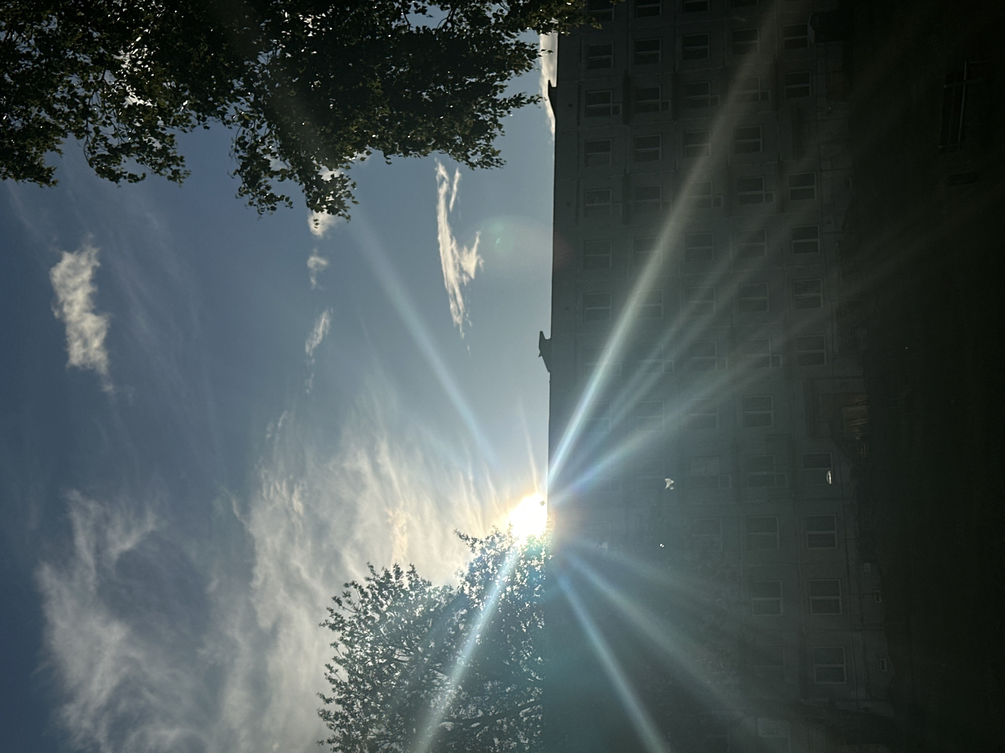 The image depicts a view of a multi-story building, possibly an apartment complex, with a partially constructed facade. The sun is setting or rising, creating a strong lens flare that radiates across the sky and illuminates the building. Silhouetted trees frame the scene on the left and right. The sky is blue with wispy clouds. A portion of a park bench is visible in the foreground. The scene is likely located in Liepaja, Latvia, given the architectural style and vegetation. There are no people or animals visible in the frame.