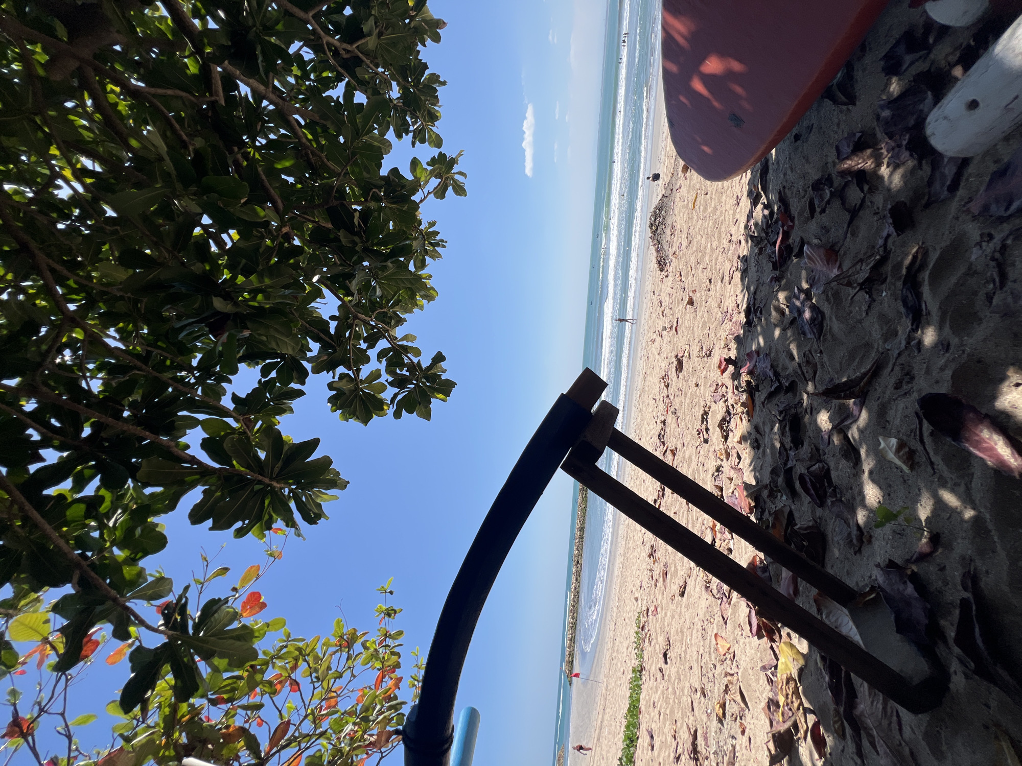 The image depicts a beach scene in Kuta, Indonesia, under a bright blue sky. In the foreground, dried leaves are scattered on the sand. A large, dark, curved object, possibly part of a beach chair or umbrella, is prominently positioned, casting a shadow. To the right, a portion of a red surfboard is visible. In the mid-ground, the sandy beach slopes down to the ocean. Several people can be seen walking along the shore and in the water, with small boats dotting the horizon. A breakwater made of rocks extends into the sea. Overhead, lush green leaves and some reddish foliage from a tree canopy frame the top of the image. The overall atmosphere suggests a sunny day at a tropical beach.