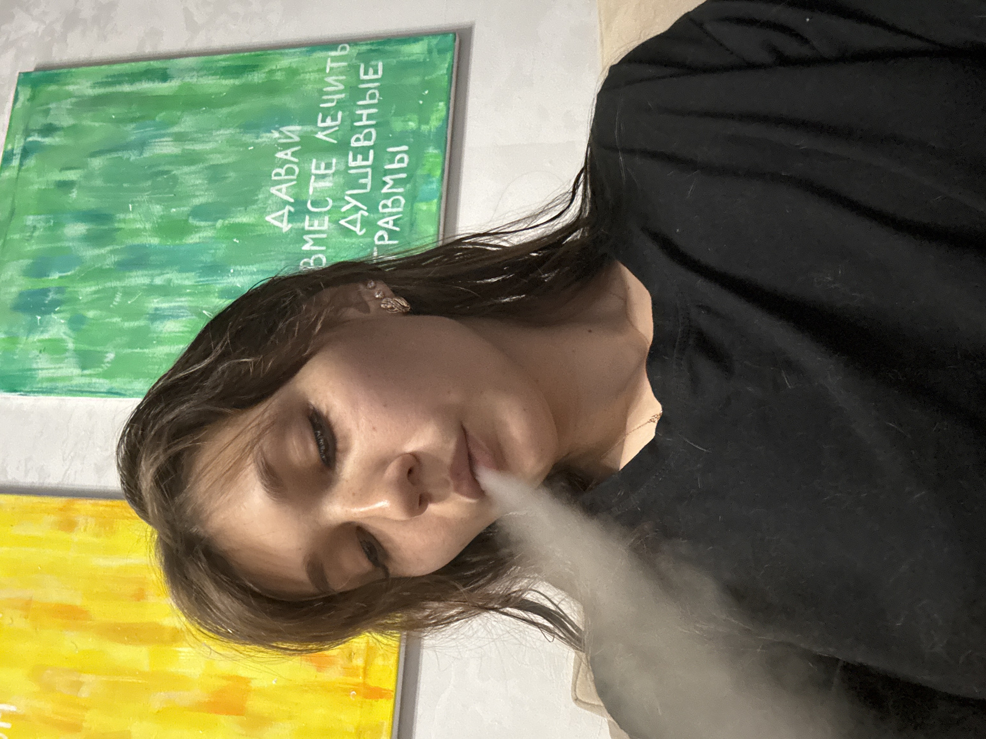 A young woman with dark hair exhales smoke, creating a cloud in front of her face. She is wearing a black t-shirt and a necklace. She has earrings in her ears. Behind her, a large green canvas with white Russian text is visible, as well as a yellow canvas on the left. The environment appears to be indoors, possibly a living room or studio. The text on the green canvas reads "ДАВАЙ ВМЕСТЕ ЛЕЧИТЬ ДУШЕВНЫЕ ТРАВМЫ" which translates to "LET'S HEAL SOUL WOUNDS TOGETHER". The image does not provide enough information to determine its exact location within Novokuznetsk, Russia.