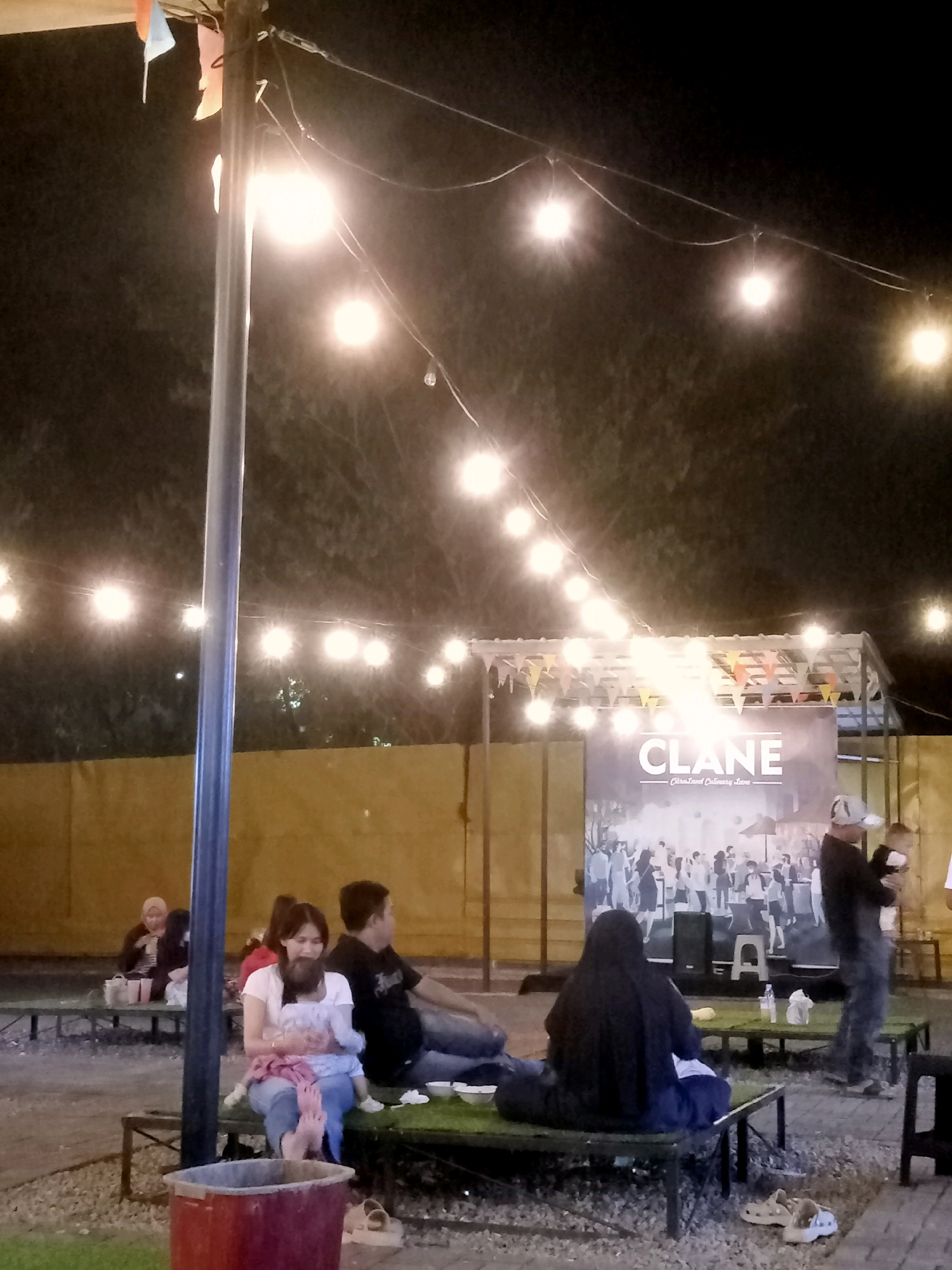 The image captures an outdoor gathering at night, likely at a food court or a similar public space in Cirebon, Indonesia. A prominent banner displaying "CLANE - Citraland Culinary Area" is visible, indicating the location. String lights illuminate the scene, casting a warm glow over the patrons seated at various tables. In the foreground, a woman is holding a baby, while other individuals are seen conversing and eating. The overall atmosphere appears relaxed and social.