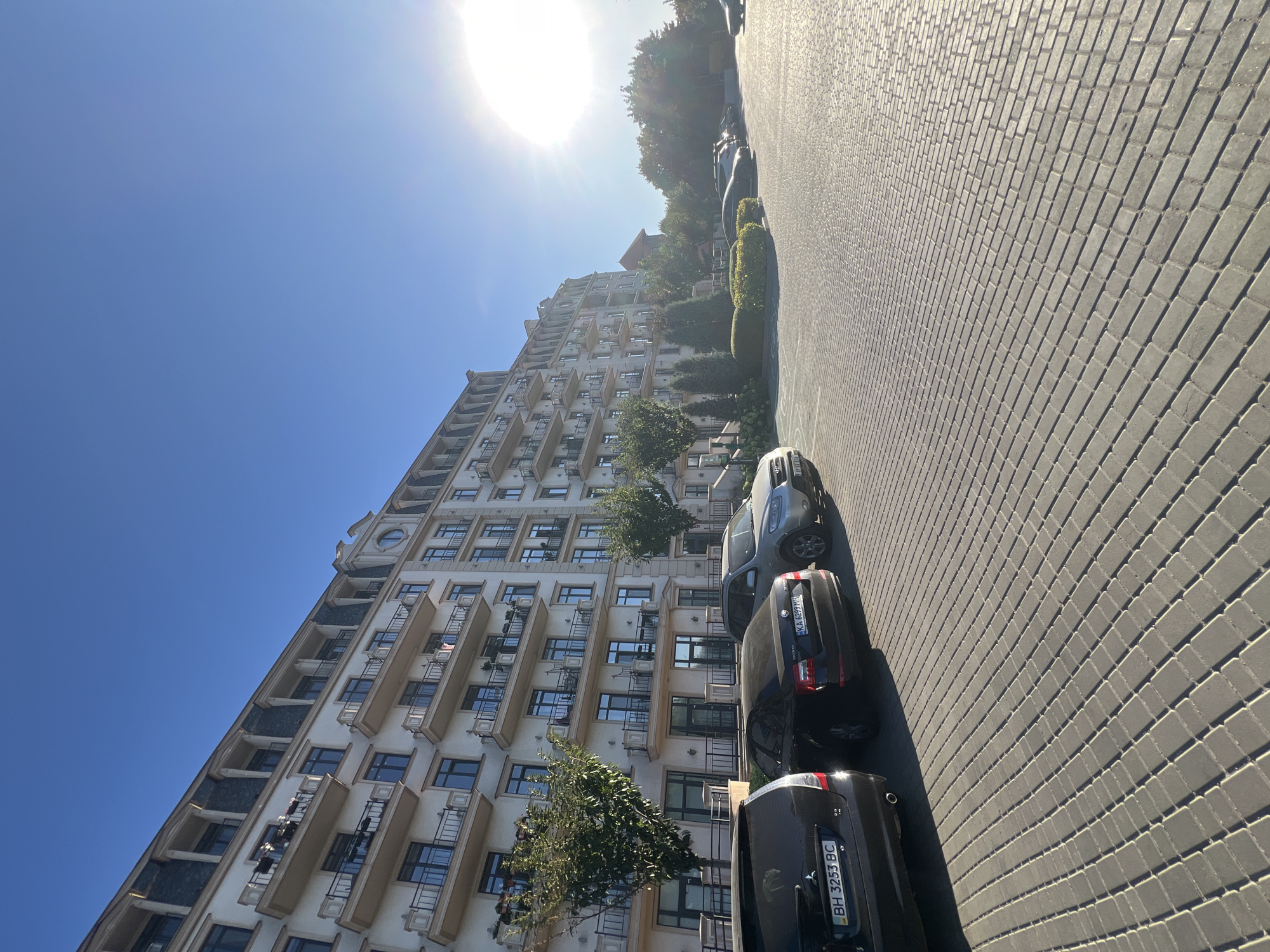 The image shows a sunlit exterior view of a modern, multi-story apartment building with a light-colored facade, multiple balconies with black railings, and a dark mansard roof. In the foreground, a paved parking lot made of rectangular gray pavers is visible. Several cars are parked in the lot, including a dark SUV on the left with license plate BH 3253 BC, a black Skoda in the middle with license plate KA 6299 MH, and a silver SUV partially visible on the right. Green trees and manicured bushes are planted along the building and in front of it. The sky is clear and blue with a bright sun in the upper right quadrant, casting long shadows across the parking lot. The setting appears to be a residential area in Fontanka, Ukraine, during daylight hours. There are no people or discernible actions taking place.
