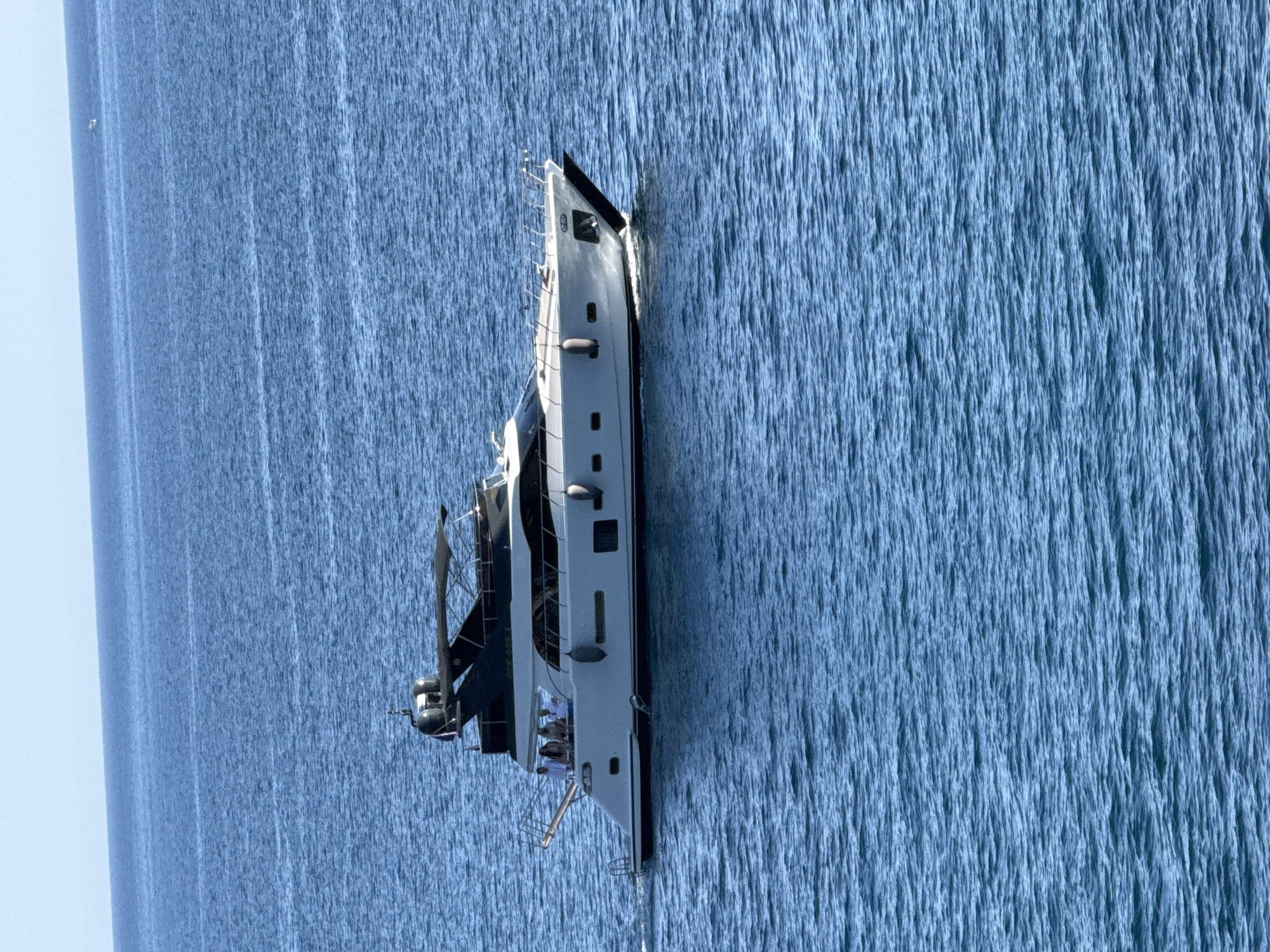 A white and black motor yacht is sailing on a clear blue sea under a bright, hazy sky. The yacht has multiple levels and a black awning. Four individuals are visible on the upper deck, appearing to be passengers. The yacht is moving from left to right, creating a slight wake. The water is calm with small ripples. In the distance, a faint, small object, possibly another boat, can be seen on the horizon. The scene is likely near the coast of Kamenovo, Montenegro, given the typical clear blue waters and boat traffic in the Adriatic Sea.