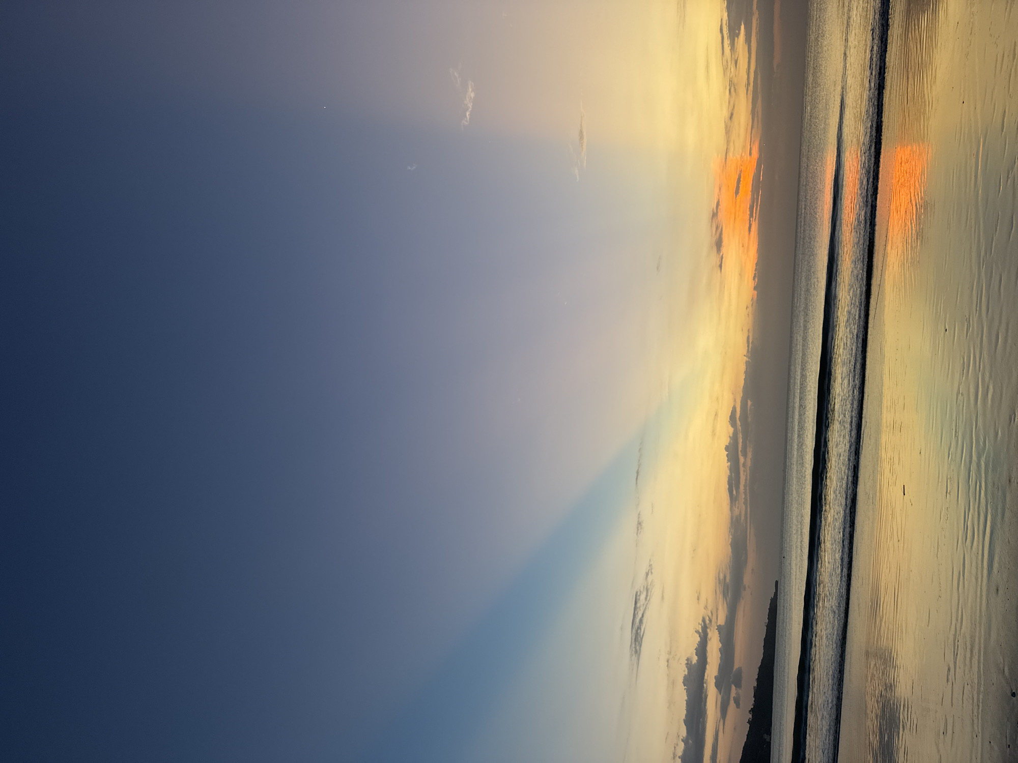 This is a photograph of a sunset over the ocean. The sky is a gradient of deep blue to bright orange and yellow, with some wispy clouds. Sunbeams are visible, fanning out from the horizon. The ocean is calm, with gentle waves lapping at the shore. The wet sand reflects the colors of the sky. The scene evokes a sense of peace and tranquility. The time of day is sunset, and the weather appears to be clear. The location is Kuta Selatan, Indonesia. There is no visible text in the image.