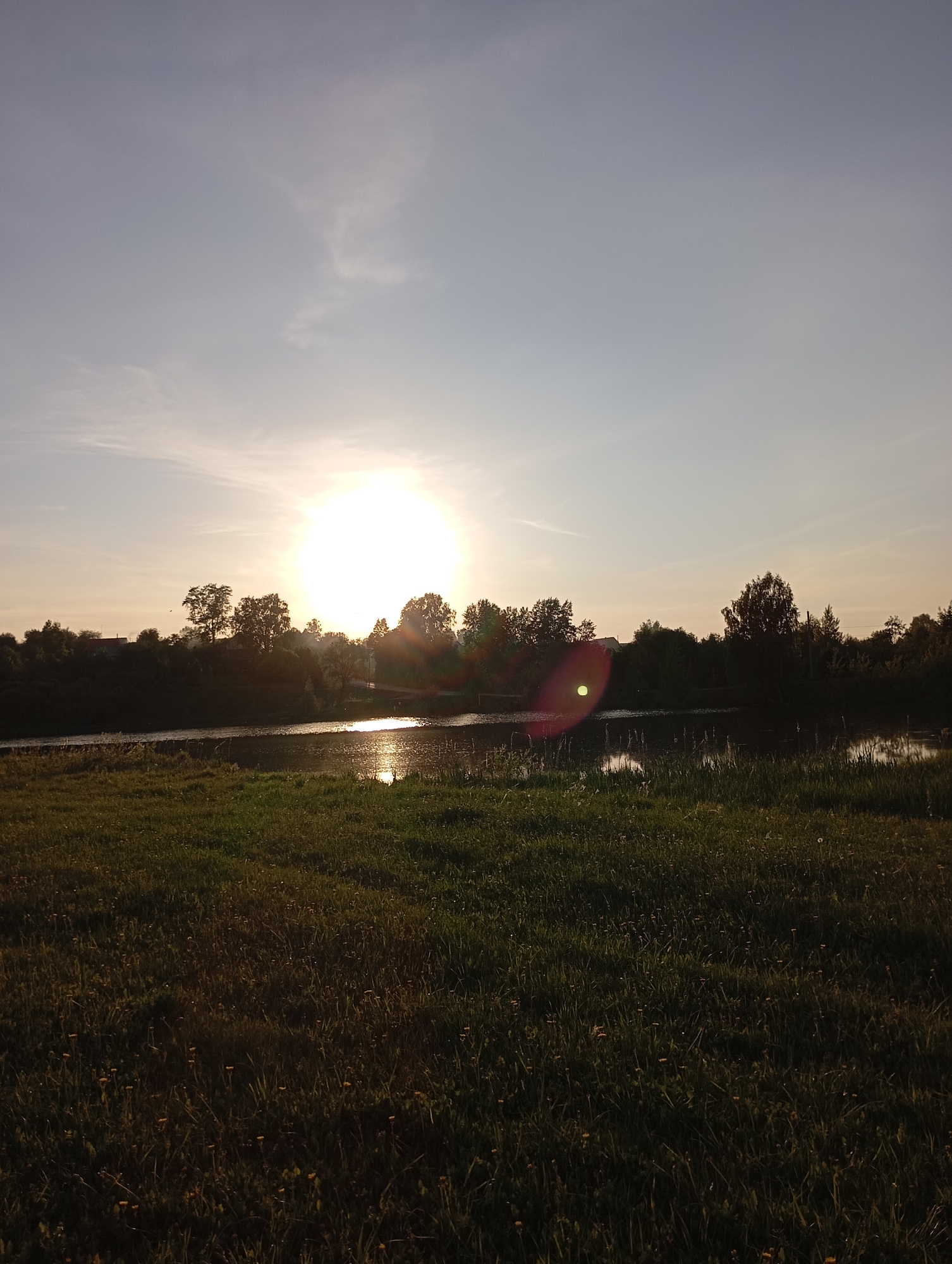 The image shows a landscape at sunset, likely in a rural area. The sun is bright and positioned behind a treeline, causing lens flare. A body of water reflects sunlight. The foreground is dominated by a field of grass. The sky is light with some cloud cover.
The location is unknown.