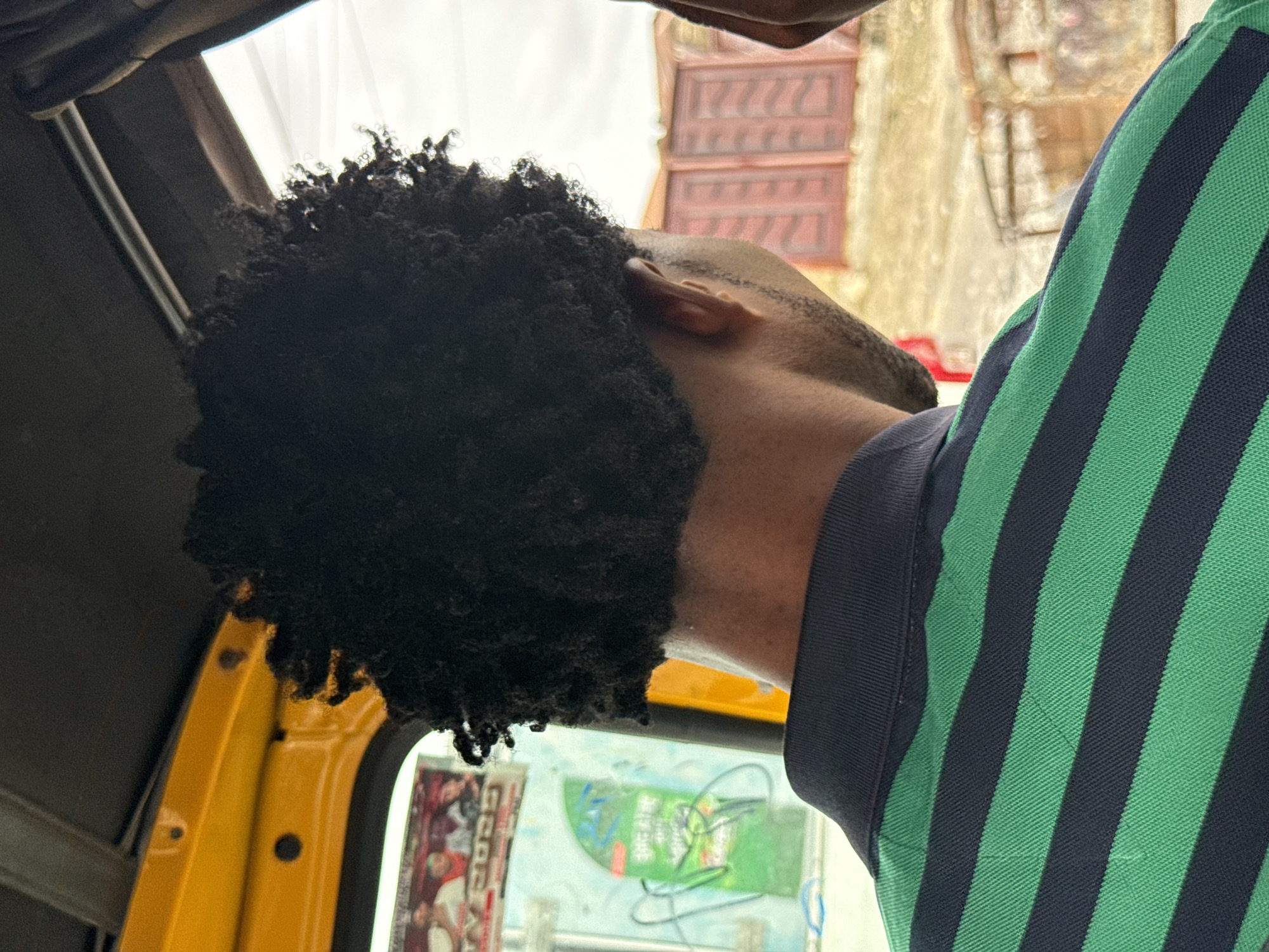 A man with short, dark, tightly curled hair, wearing a striped green and black polo shirt, is inside a yellow vehicle, likely a "Keke" or auto rickshaw, in Ado, Nigeria. The view is from inside the vehicle, looking towards the passenger. Part of the vehicle's frame is visible, as is a window with stickers on it. Outside the vehicle, the background is blurred, but shows a building with stacked red shutters.
