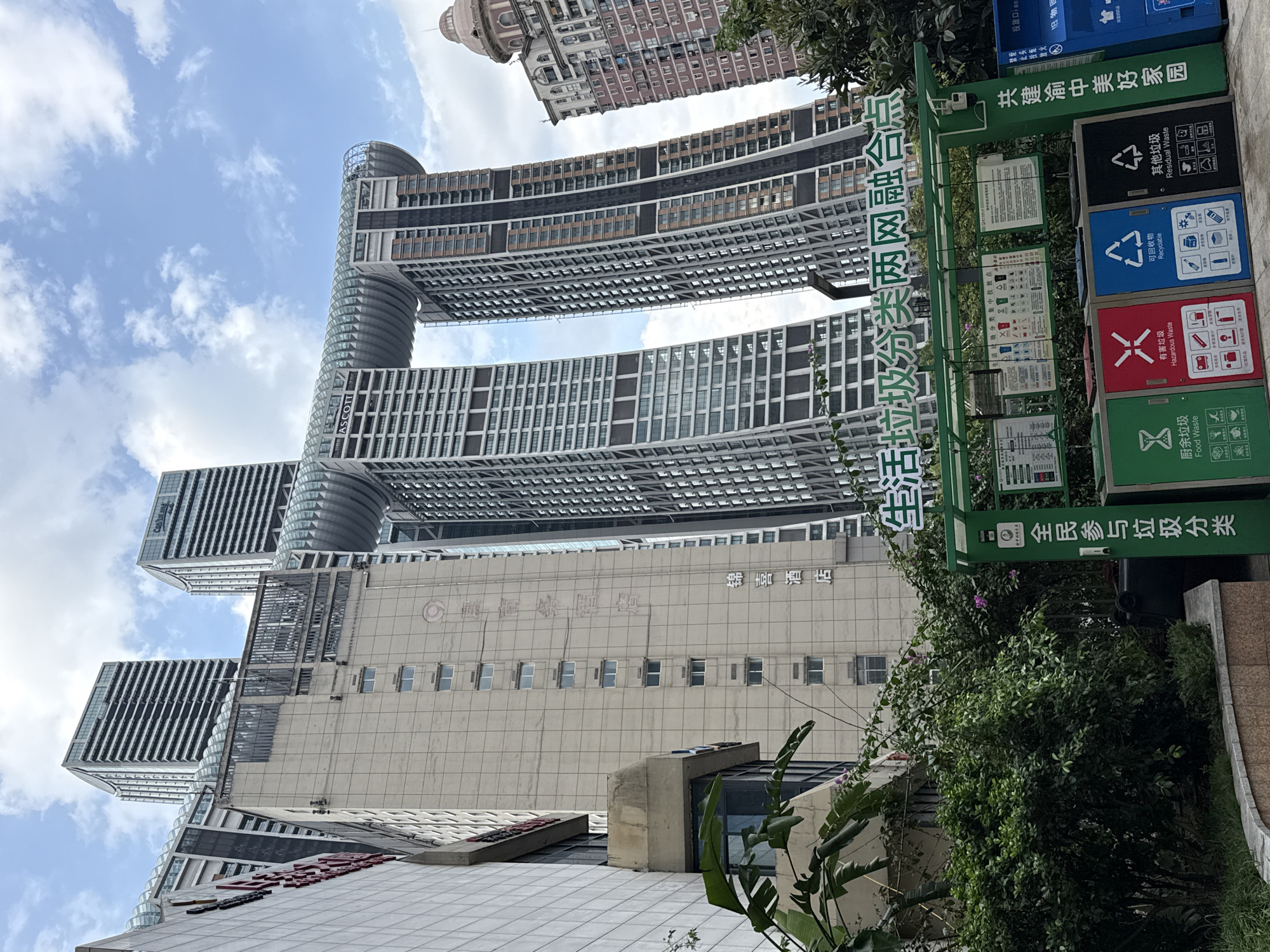 This is a medium-shot, eye-level, outdoor, daytime, vertical photograph taken in the Yuzhong District of China. The image features several towering, modern skyscrapers with distinctive architectural designs. The most prominent structure is a building with two arched sections that connect two sets of towers, creating a unique, sky-bridge-like appearance. The building on the left appears to be a hotel, with the word "ASCOTT" visible on its facade. The foreground is dominated by lush green foliage and a set of brightly colored, modern recycling bins. The bins are clearly labeled with icons and text in Chinese, indicating different categories of waste such as "Food Waste," "Hazardous Waste," "Recyclable," and "Residual Waste." Above the bins, a green sign with Chinese text is mounted, likely providing information about waste sorting. The sky is partly cloudy with a mix of blue sky and white clouds. The overall scene suggests an urban environment focused on modern architecture and public facilities like waste management.