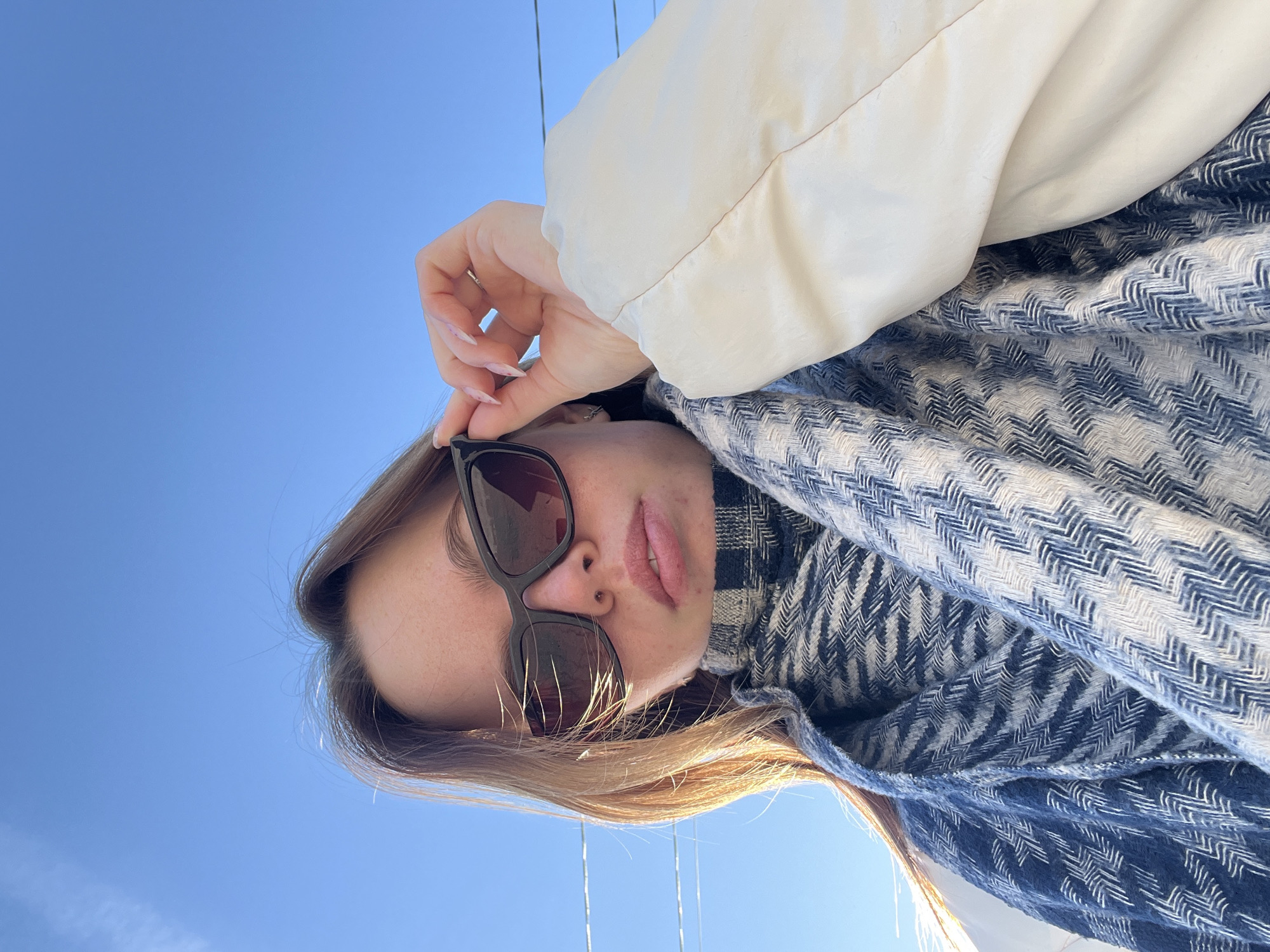 A woman is depicted in a low-angle shot against a clear blue sky, likely taken outdoors in Chaikovsky, Russia, though this location is not explicitly verifiable from the image itself. She is wearing sunglasses and has fair skin. She's partially wrapped in a blue and white patterned scarf and appears to be wearing a light-colored coat or jacket. Her right hand is near the top of her sunglasses, and her head is slightly tilted to the right in the frame. Her expression is neutral and lips are lightly visible, as if parted. Her light brown hair is visible. The scene is bathed in natural light.