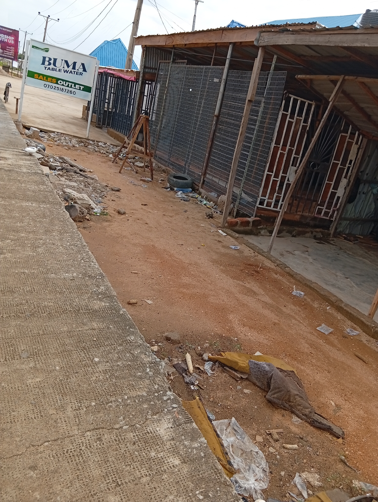 The image depicts an outdoor scene in Jalingo, Nigeria, likely a roadside commercial area. The foreground is dominated by a cracked, textured concrete pathway. To the right of the pathway, the ground transitions to reddish-brown dirt, littered with debris including plastic bags, discarded items, and what appears to be a partially covered, elongated object.
Further into the scene, a makeshift structure is visible, characterized by a corrugated metal roof supported by wooden posts and beams. The structure has an open front, partially covered with wire mesh fencing and metal bars. Behind this structure, a blue, angled roof can be seen, possibly part of a different building.
A prominent white sign with green and blue lettering stands to the left of the scene. The text on the sign reads: "BUMA TABLE WATER" and below that, "SALES OUTLET" followed by a phone number: "07025187260". The overall impression is one of a developing or perhaps neglected commercial space. The lighting suggests it is daytime, with overcast or diffused light, indicating potentially cloudy weather. No people are visible in the frame. The scene conveys a sense of everyday commerce and the urban environment in Jalingo.