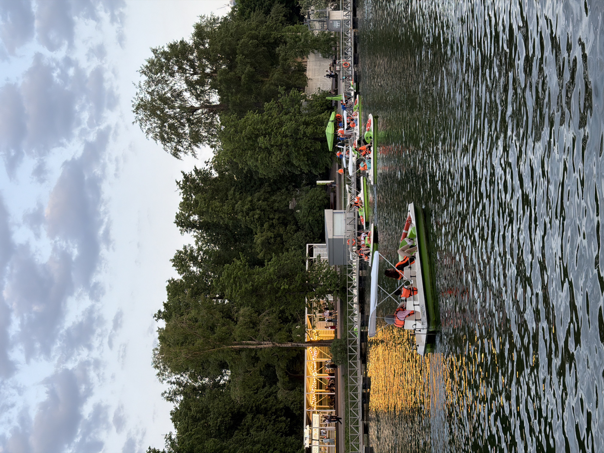 The image shows a waterfront scene, likely in Moscow, Russia. The water surface reflects the golden light of the setting sun. Numerous paddle boats and kayaks are docked along a pier, each occupied by individuals wearing orange life vests. On the shore, a structure decorated with string lights suggests a restaurant or entertainment venue. Groups of people are visible along the pier and within the structure. Tall, mature trees line the waterfront, and the sky above is covered with light clouds.