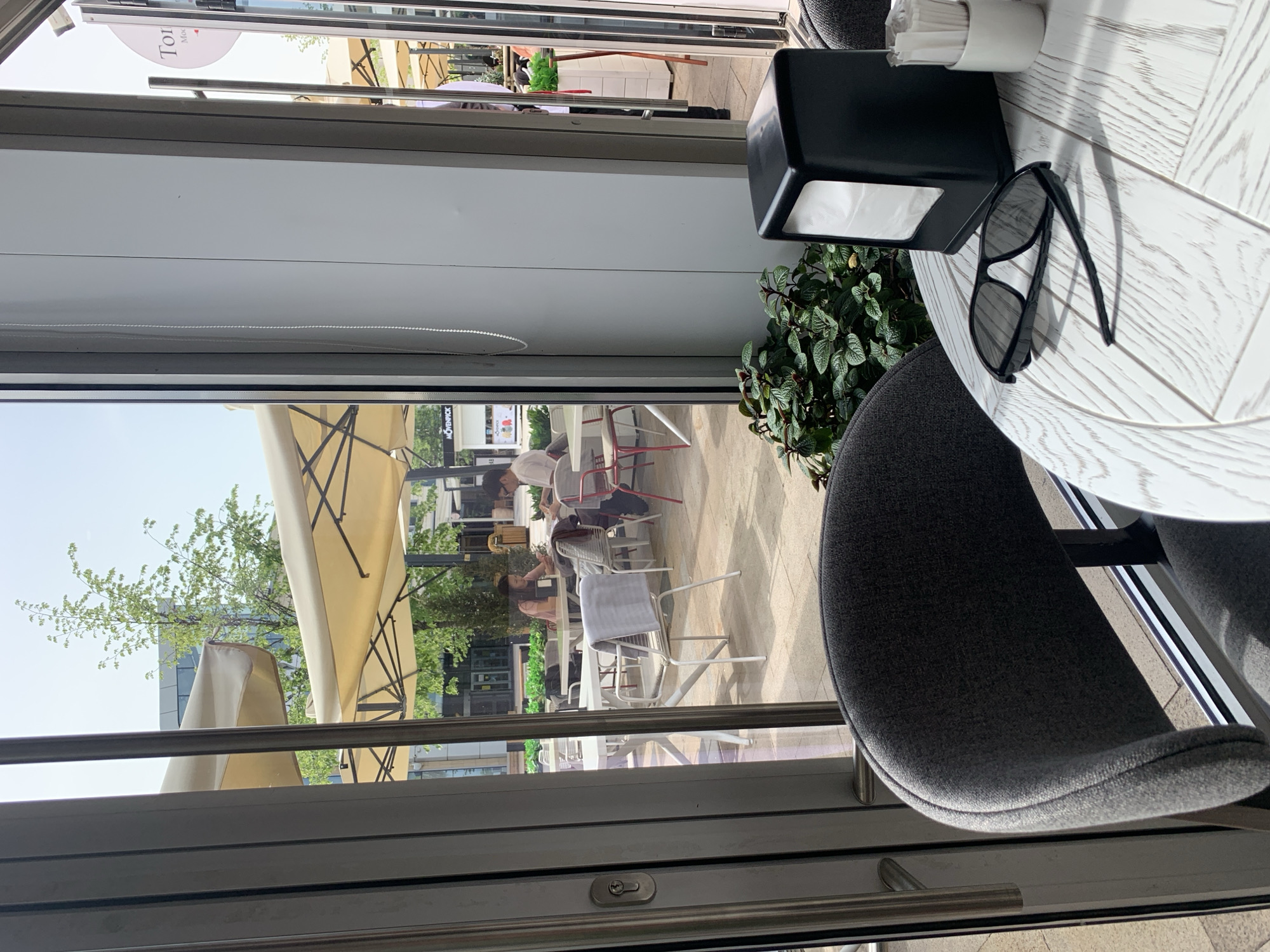 The image shows a view from inside a cafe in Almaty, Kazakhstan, looking out onto an outdoor seating area. A partially visible gray chair occupies the foreground along with a round white table with a black napkin holder, a container of white napkins, a small plant, and a pair of black sunglasses. The cafe's glass doors offer a view of patrons seated at tables outside under large beige umbrellas. People are engaged in conversation and dining. Trees are visible in the background, along with building architecture consistent with a modern urban environment. The daylight indicates it is daytime.
