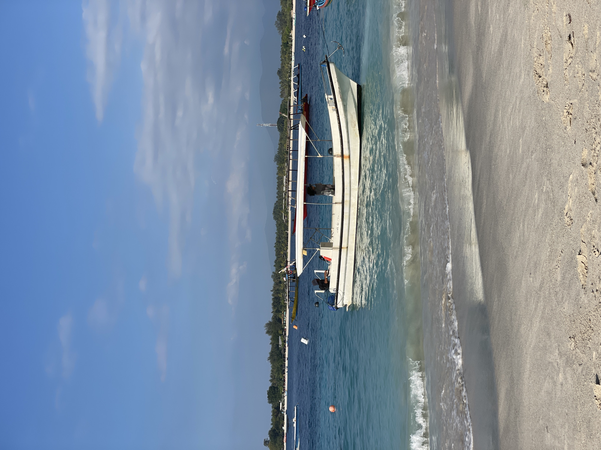 The image depicts a beach scene in Indonesia, likely on an island given the boats and clear blue water. In the foreground, a sandy beach meets gentle waves. Several boats are visible in the turquoise water. The closest boat is a white vessel with a red-trimmed canopy, a person appears to be steering it from the rear. Further out, other boats, including a yellow one and a longer white boat with a canopy, are anchored. In the background, a treeline suggests a coastal area with vegetation, and beyond that, mountains are faintly visible under a sky with scattered clouds. The overall impression is of a tranquil tropical setting.