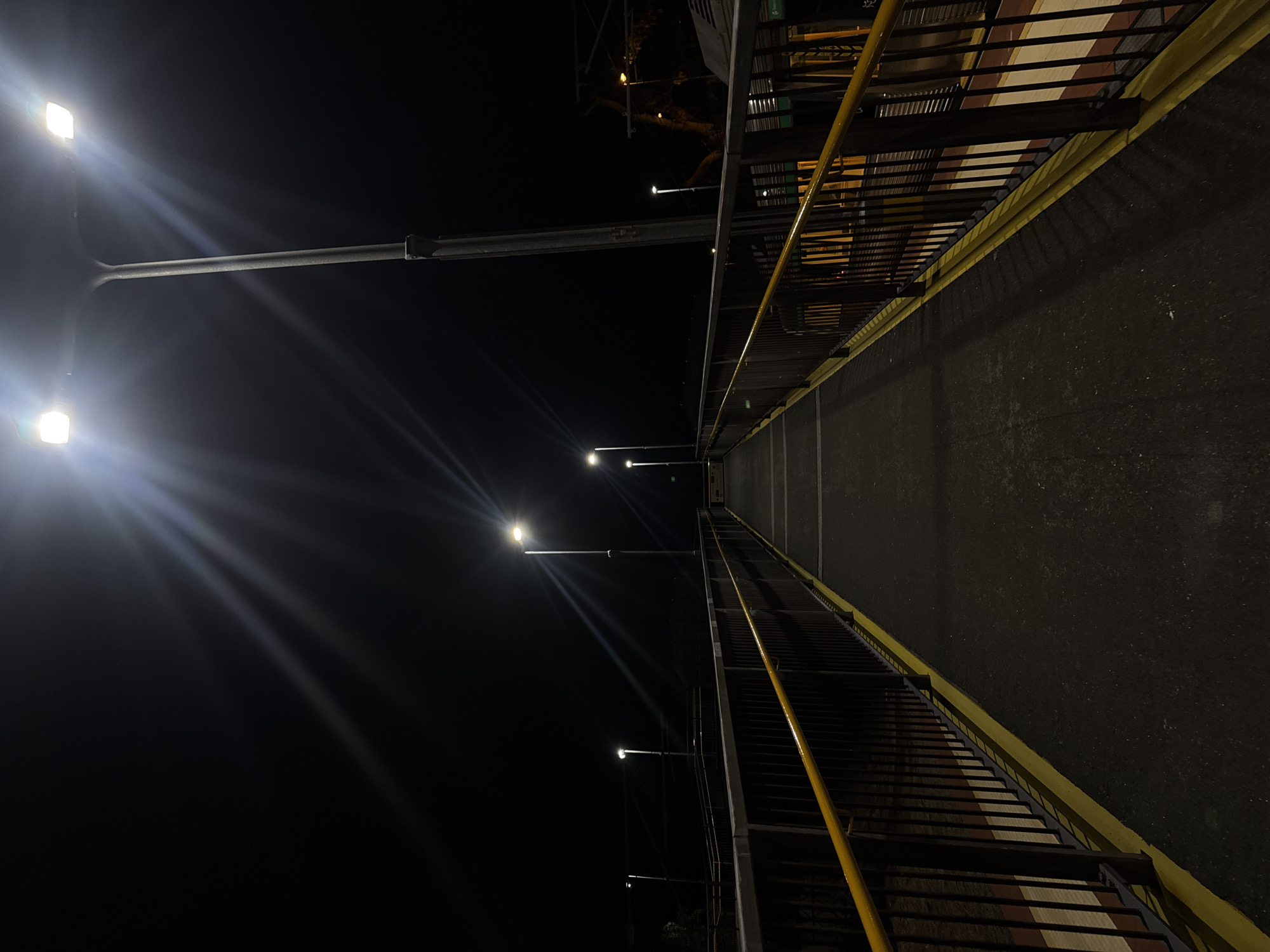 The image shows an elevated pedestrian walkway at night in Bassendean, Australia. The walkway is flanked by dark railings on both sides, painted yellow on the inner part. Streetlights illuminate the walkway. In the distance the path appears dark and the view is straight along the path.