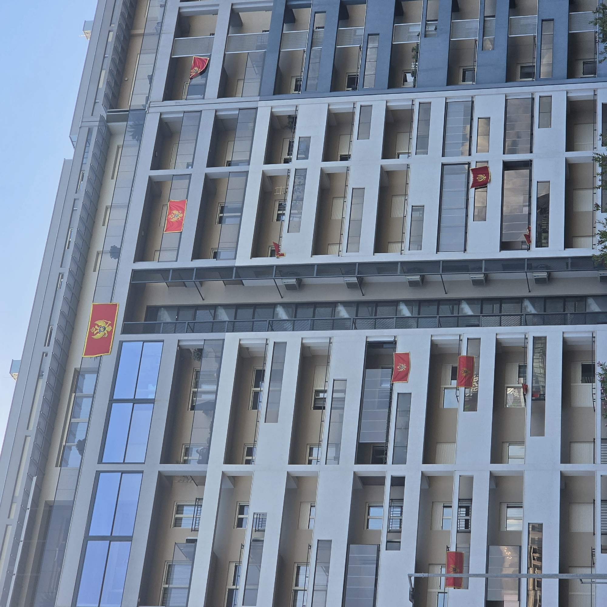 The image captures the facade of a tall, modern building in Podgorica, Montenegro, adorned with multiple Montenegrin flags hanging from balconies. The building has a grid-like architectural design with alternating sections of glass and solid panels. The flags are predominantly red with the Montenegrin coat of arms. The sky is visible in the upper left corner.