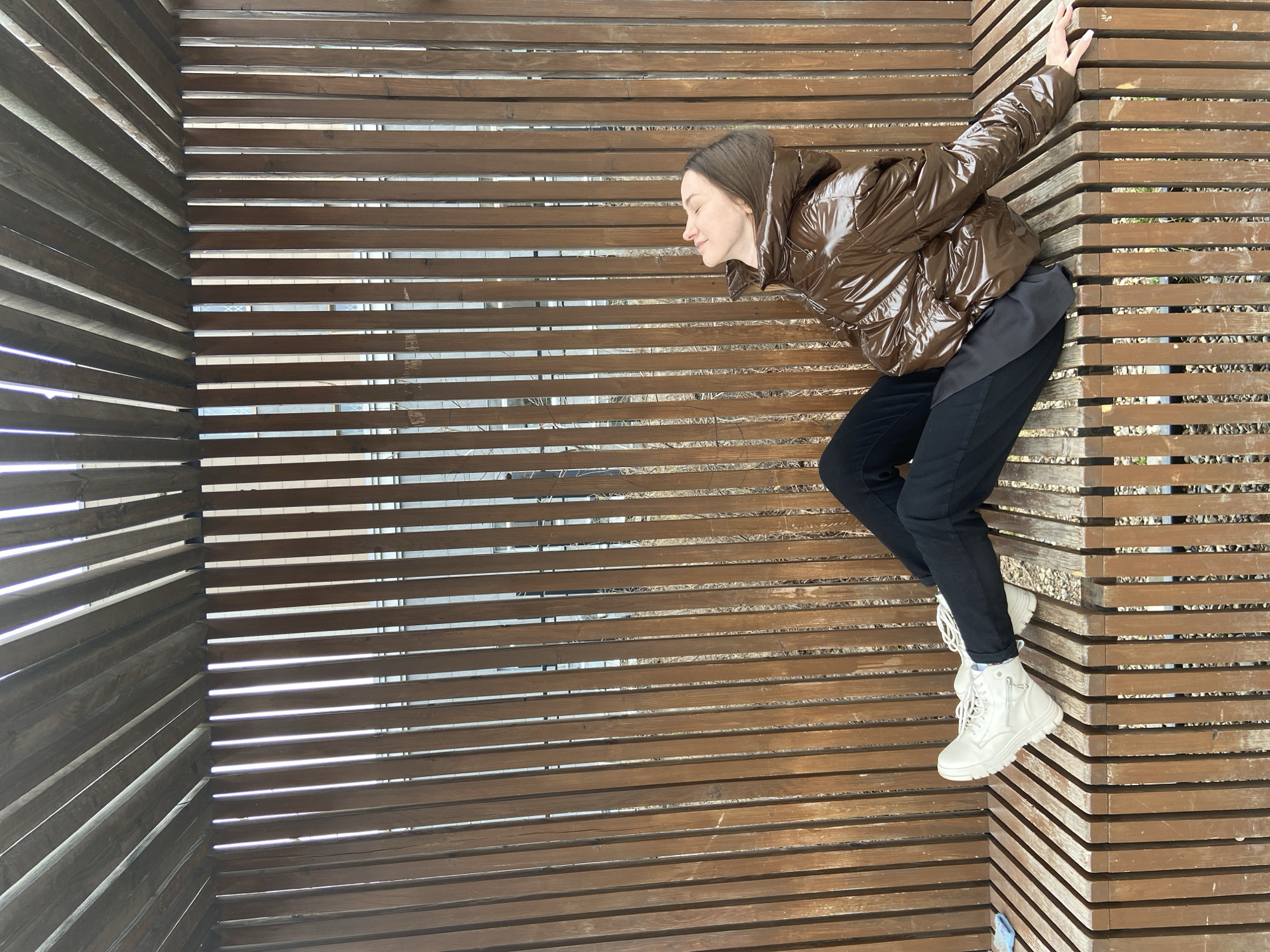 A woman is propped up against a wooden structure with horizontal slats in Izhevsk, Russia. She wears a shiny brown puffer jacket, black pants, and white combat boots. Her eyes are closed, and she appears to be relaxing. The structure consists of closely spaced, uniform wooden slats, oriented horizontally, creating a linear pattern. The slats form a corner behind her, extending both to her left and right. Sunlight filters through the gaps between the slats. The ground beneath the structure is visible through the gaps between the slats, showing gravel or small stones.