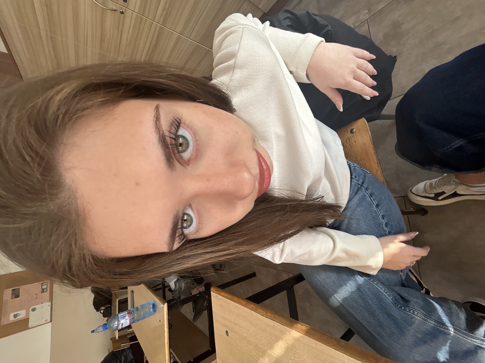 The image depicts a young woman in an indoor, educational setting, likely a classroom or lecture hall, in Sofia, Bulgaria. She is positioned close to the camera, looking directly forward with wide green eyes, her face dominating the upper portion of the frame due to a low-angle, wide-lens perspective. Her brown hair is parted on the side and falls over her shoulders. She wears a white, ribbed long-sleeve top and blue denim jeans. A silver-toned ring is visible on the ring finger of her left hand, which rests on her leg. Her fingernails have French tips.
The woman is seated on a chair with a wooden seat and metal frame. In the background, several light wooden desks with metal frames and matching chairs are arranged in rows. On a desk to the left, a partially consumed plastic bottle of "DEVIN" mineral water is visible. Further back, a dark bag or backpack rests on a chair. The wall behind the desks features a cork board with various papers pinned to it, including what appears to be diagrams or notices. Another wall section on the right exhibits light brown wooden paneling. The floor consists of light-colored tiles. The overall environment suggests an active learning space within an educational institution in Sofia, Bulgaria, supported by the presence of the Bulgarian "DEVIN" water brand.