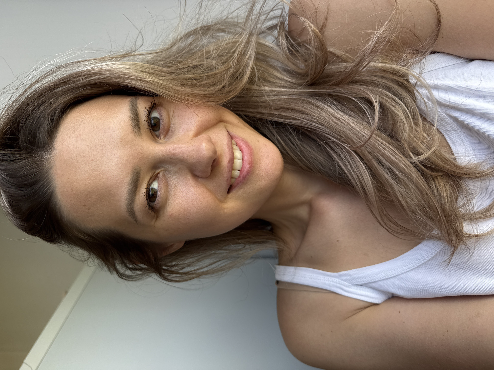 This media file is a close-up, portrait-style photograph of a woman. The image is oriented sideways, with the woman's head tilted to the right. She has medium-length, wavy blonde hair with darker roots, which falls around her face and over her shoulder. Her eyes are brown and she is smiling, revealing straight, white teeth. She is wearing a white tank top.
The background appears to be a plain, light-colored wall, suggesting an indoor setting. The lighting is soft and natural, hinting at daytime. The woman's expression is friendly and relaxed, conveying a sense of joy or amusement. There are no visible objects or text within the frame, making the woman the sole focus of the image. The overall impression is casual and personal.